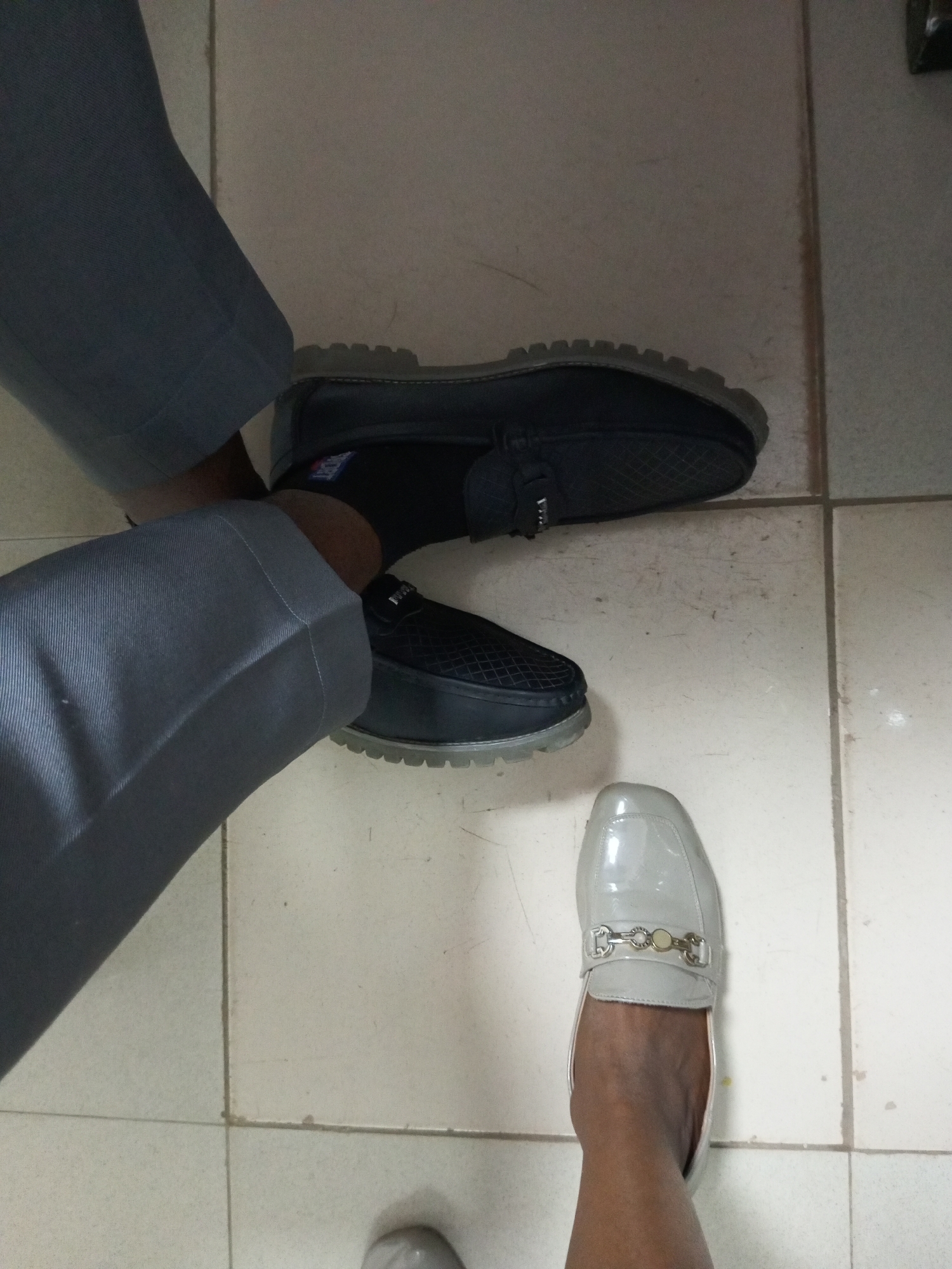 This is a close-up, top-down shot of two people's legs and feet on a tiled floor, likely indoors in Bukuru, Nigeria. The person on the left is wearing grey trousers and black, slip-on shoes with a textured pattern and clear soles. This person also has dark socks visible. The person on the right is wearing grey trousers and a light grey, patent leather loafer with a decorative silver buckle. The tiled floor appears to be light-colored with visible grout lines. The lighting suggests it could be daytime, possibly near a window or under artificial light.