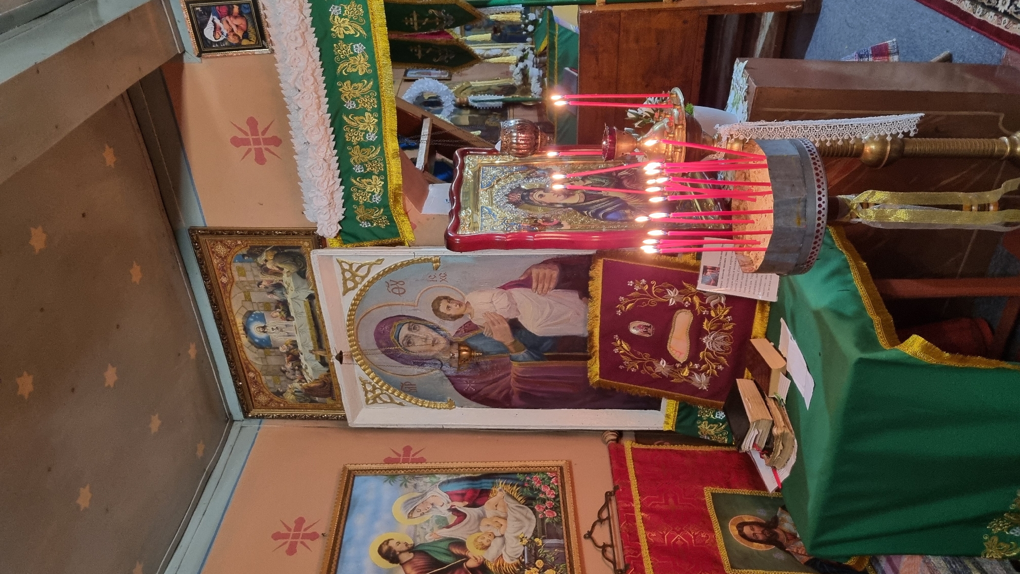 This image depicts the interior of an Eastern Orthodox church in Vyshka, Ukraine, possibly during a religious service or a moment of prayer. The scene is rich with religious iconography and ceremonial objects.
Dominating the foreground are several icons, including a large, framed painting of the Virgin Mary holding the Christ Child. To its right, a smaller icon shows the same subject, with numerous red candles burning in front of it, their flames casting a warm glow. A metallic candelabra holds many of these candles. Another framed icon to the left depicts a biblical scene, possibly the Last Supper. Below these, another icon shows the Holy Family.
The church's walls are painted a soft peach color and adorned with decorative star patterns and a floral motif. Green and red liturgical cloths, edged with gold trim, are draped over surfaces and along the altar area, indicating a sacred space. A wooden lectern or table is visible in the background, with books and papers placed upon it. The overall atmosphere is one of reverence and devotion. The lighting suggests it is daytime, but the exact time is not discernible. The presence of icons, candles, and liturgical vestments are clear indicators of the religious setting. There is no discernible text in the image.