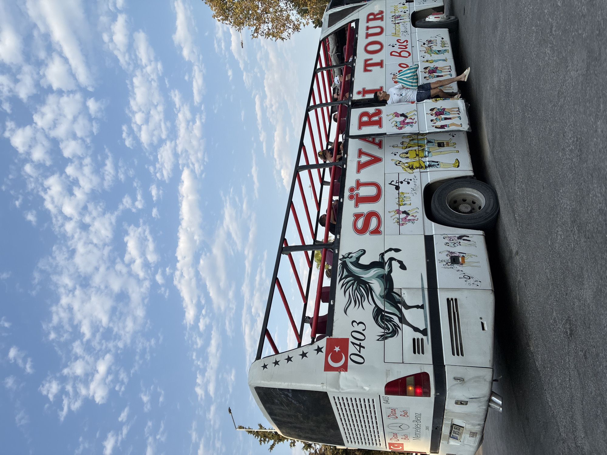 In Alanya, Turkey, a white double-decker tour bus with an open-top upper deck is parked on a paved road. The bus is decorated with colorful drawings of people dancing and music notes. A large black and white horse is depicted on the side of the bus, along with the text "SÜVARI TOUR". The Turkish flag is visible on the rear of the bus. Several passengers are seated on the upper deck, and a young man holding a striped bag is standing by the open door on the lower deck, seemingly disembarking or preparing to board. The sky above is blue with scattered white clouds. Trees and a lamppost are visible in the background.