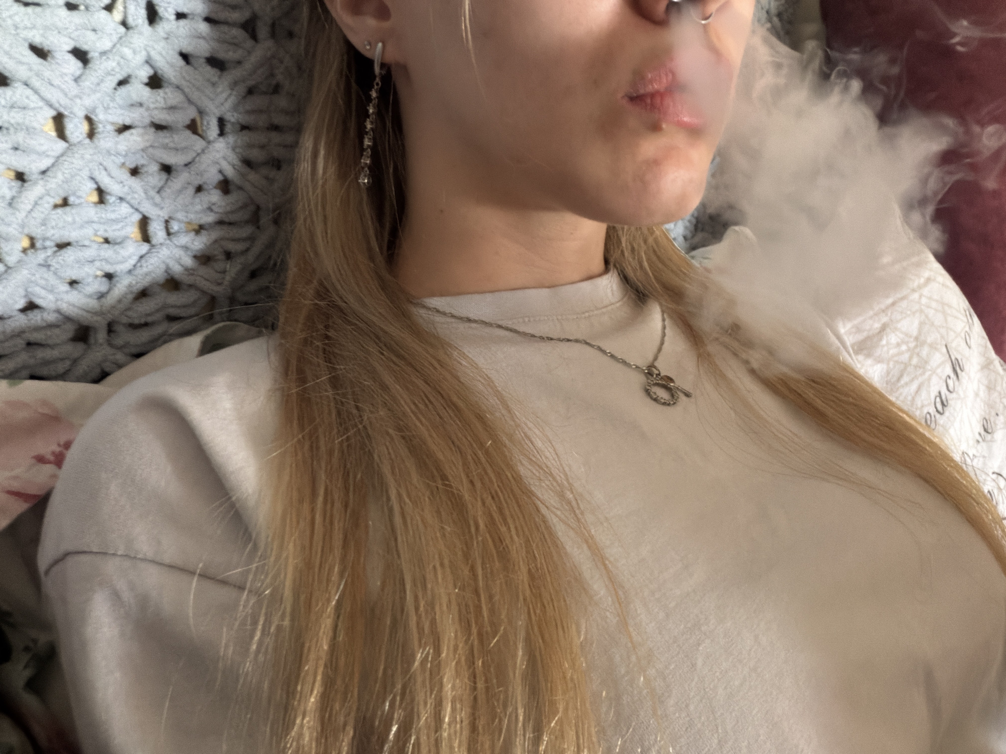 A young adult female is reclining, exhaling a cloud of smoke. She is wearing a light-colored t-shirt, a silver necklace, and a dangling earring. Her hair is blonde and long. She is lying on a bed with patterned bedding. A blue crocheted blanket is visible to her left. The location and city are unknown.
