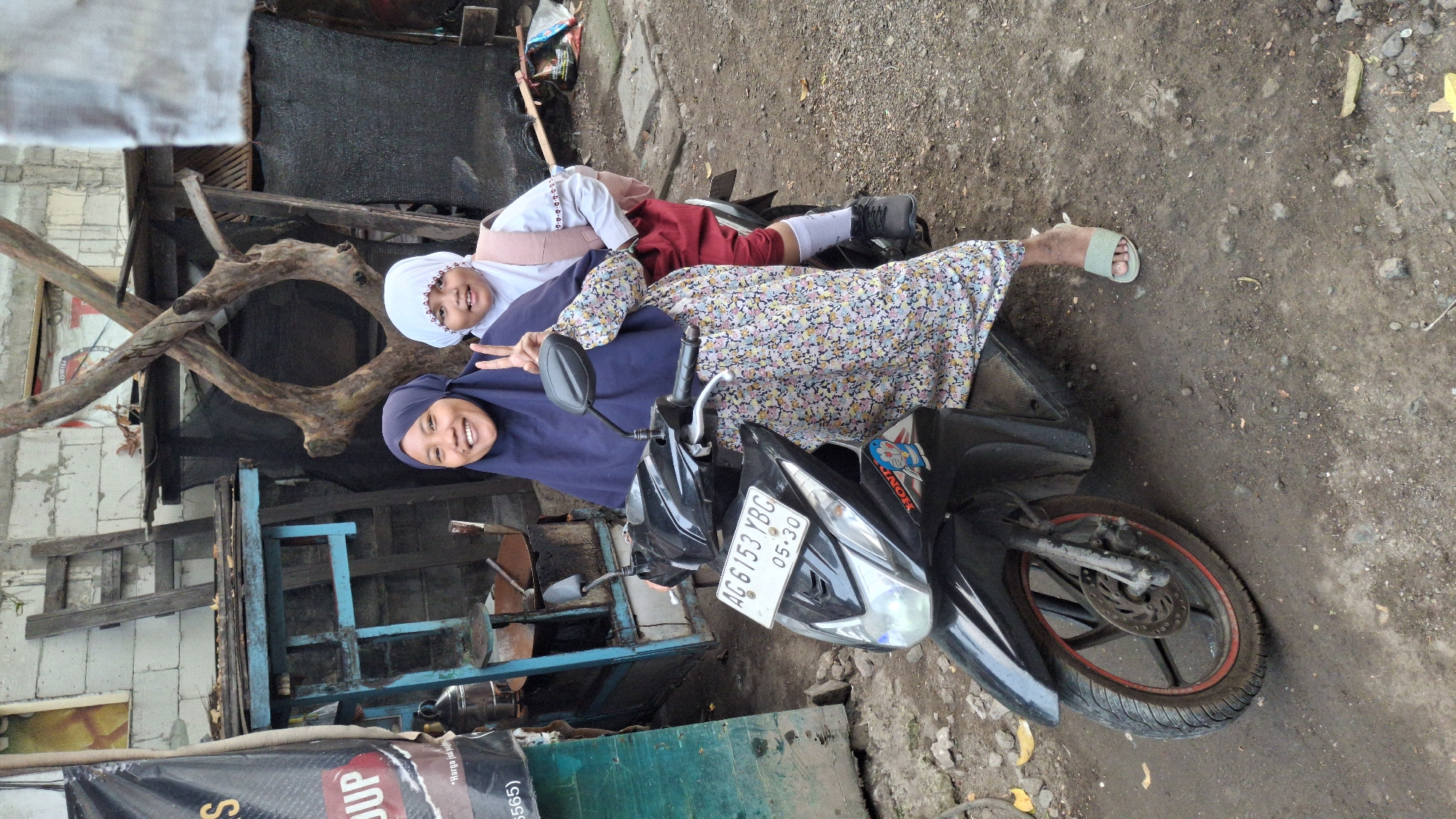 In Wadungasri, Indonesia, two individuals, likely mother and child, are posing on a black Honda scooter. The woman in front wears a dark blue hijab and a long, floral-patterned dress. She smiles widely, holding up two fingers in a peace sign. Behind her, a child in a white hijab and a light pink vest over a white shirt and red pants smiles, also holding up two fingers. The scooter's license plate reads "AG 6153 YBG" and "05-30". The background features a weathered wooden structure, a ladder, and a pile of wood, suggesting a somewhat rustic or unpaved environment. The ground is dirt and gravel.