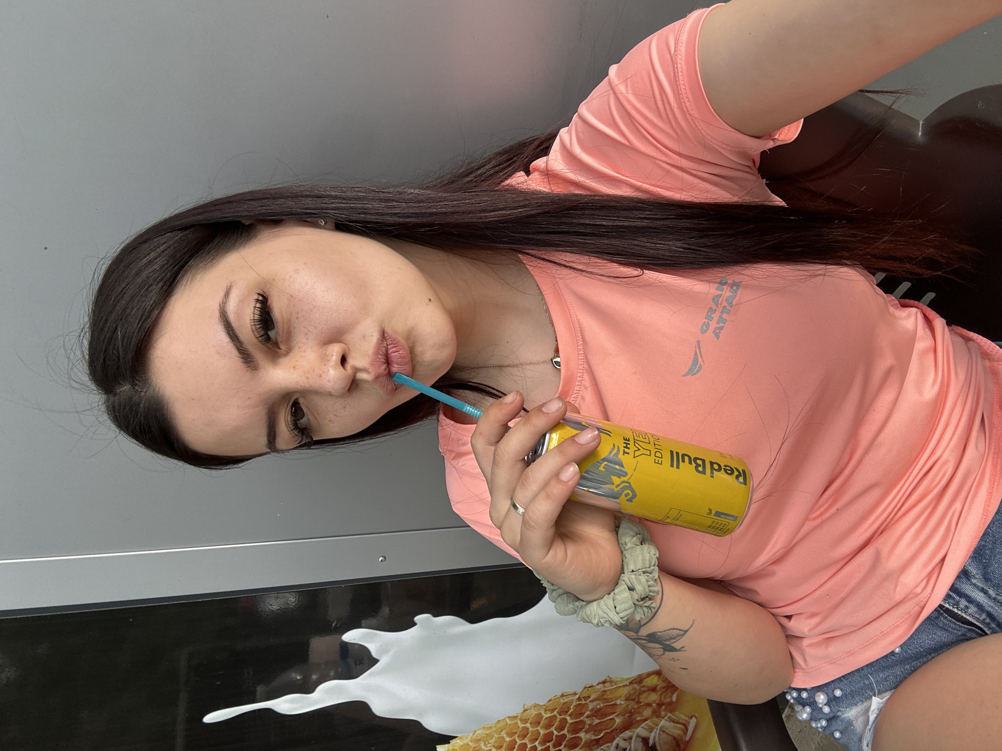 A young woman with dark hair and freckles is taking a selfie while holding a yellow can of Red Bull with a blue straw. She is wearing a coral-colored t-shirt and denim shorts adorned with pearls. The background appears to be outdoors, possibly near a building with a gray wall and a graphic design on a black surface that includes what looks like a honeycomb pattern. The lighting suggests it is daytime.