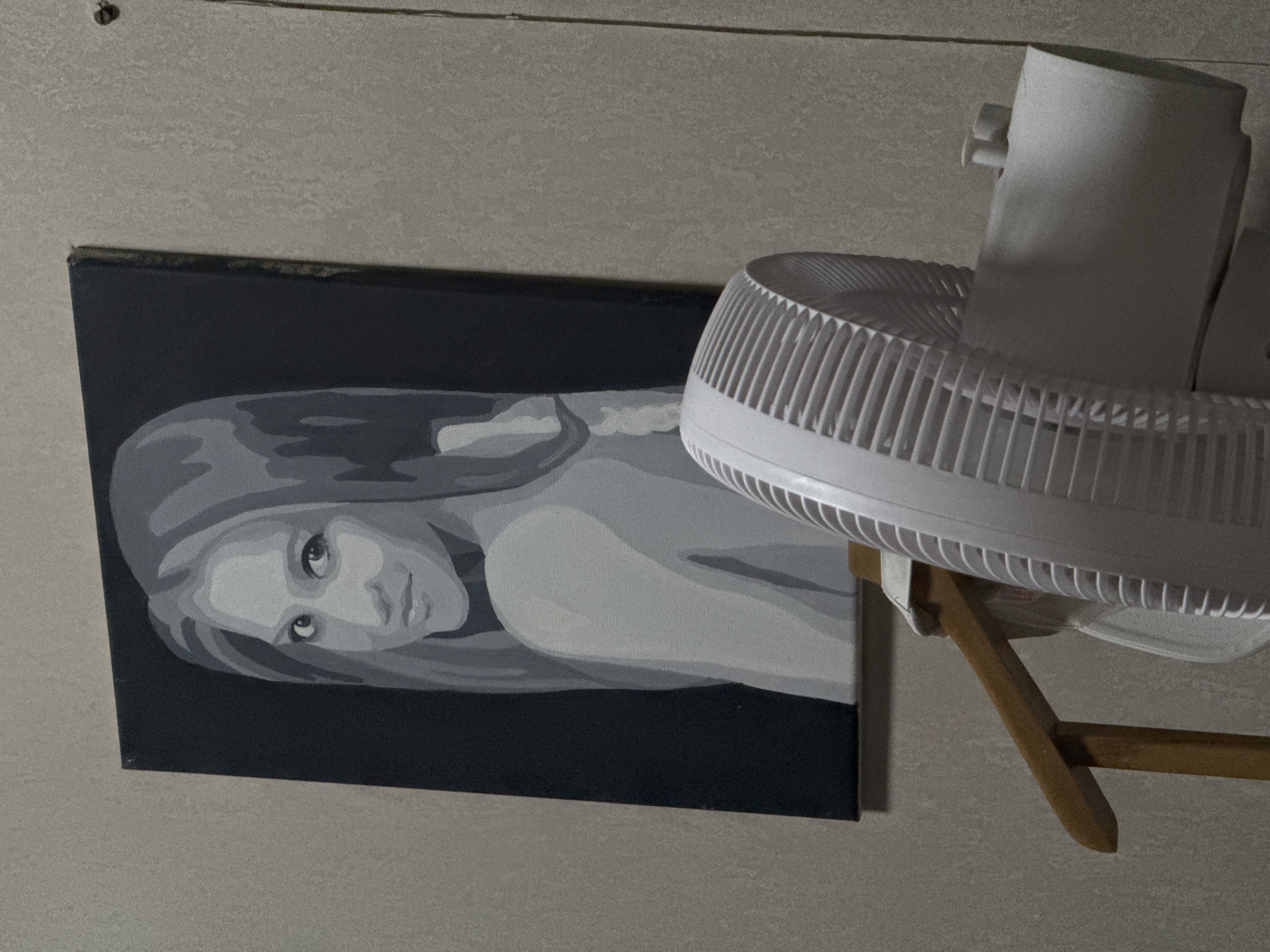 A grayscale painting of a woman's portrait is mounted on a textured, light beige wall. The woman in the painting has long, wavy hair and is looking over her shoulder towards the viewer with a subtle smile. Her shoulders and upper back are bare. In the foreground, a white electric fan with a protective grill is partially visible, positioned below and to the right of the painting. A wooden chair arm is also visible between the painting and the fan. The scene appears to be indoors, likely a room in a residential or studio setting. There is no information in the image to suggest its location in Izhevsk, Russia, or any specific city.