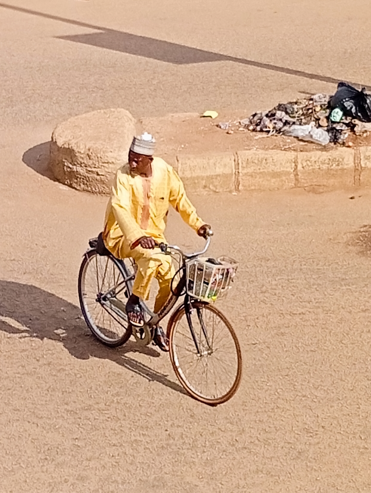 A man in a yellow traditional outfit and a patterned cap rides a bicycle on a dusty, light brown road. The bicycle has a front basket containing various items, and the man is wearing dark sandals. Behind him, to the right, is a pile of trash, including plastic bags and scattered debris, on a raised concrete curb. To the left of the man, there is a large, light brown, rounded rock formation. A long, dark shadow is cast across the road from an unseen object, likely a structure or tree, from the upper left of the frame. The scene appears to be in an outdoor urban environment in Bukuru, Nigeria, characterized by arid conditions and visible refuse.