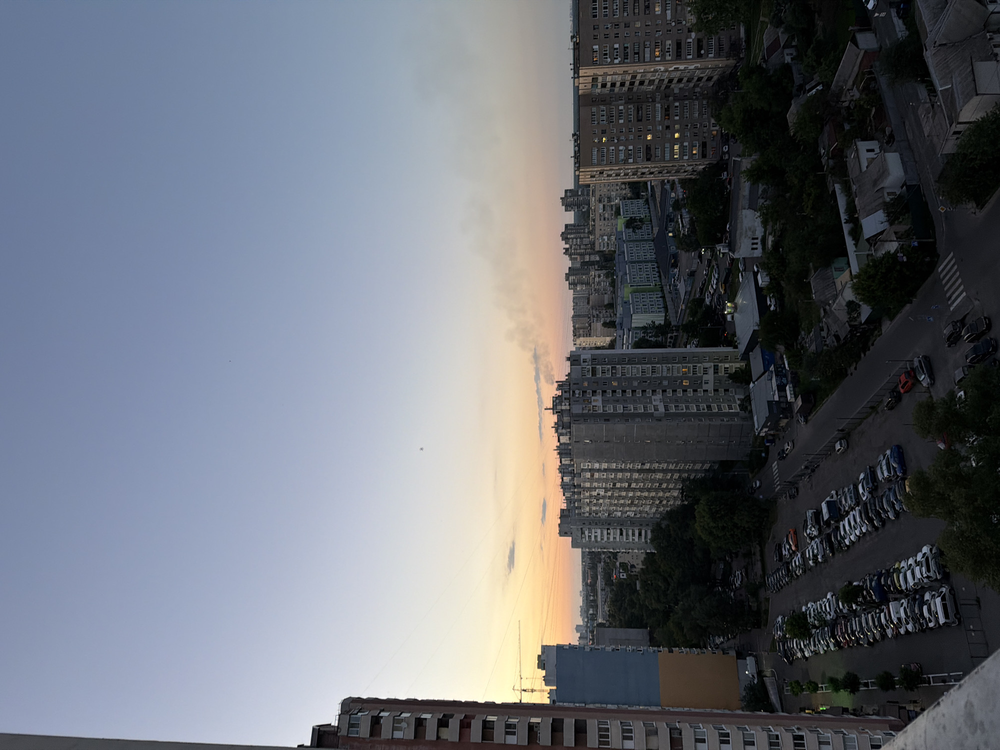 The image shows a city skyline in Kyiv, Ukraine, at dusk. In the foreground, on the left, is a tall apartment building with a reddish-brown facade. Adjacent to it is a blue and orange apartment building. To the right, a dense collection of apartment buildings of varying heights and architectural styles dominate the middle ground. These buildings have many windows, some of which are illuminated. A large parking lot filled with numerous cars is visible in front of the apartment buildings. Beyond the parking lot and apartment buildings, lush green trees are interspersed throughout the urban landscape. In the distance, the sky transitions from a pale blue to a warm orange and yellow hue near the horizon, indicating the setting sun. A faint plume of smoke is visible in the sky above the buildings on the right side of the frame. The overall scene depicts a typical residential area of Kyiv during the evening.