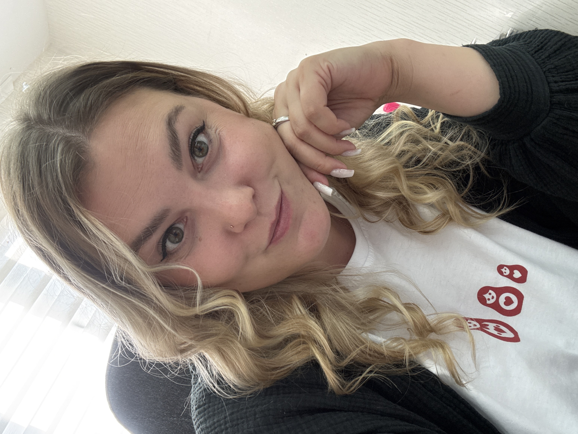 The image shows a close-up, eye-level, selfie-style shot of a Caucasian woman with blonde, wavy hair. She is wearing a white t-shirt with a red nesting doll graphic and a dark-colored cardigan. Her right hand is resting on her chin, and she is wearing a silver ring on her left ring finger and has long, white, glittery fingernails. She has a nose ring and her eye makeup is prominent. The background appears to be an indoor setting with textured white walls and possibly a window with blinds on the left side. The lighting suggests natural light coming from the left. The woman is looking directly at the camera with a slight smile. There is no information in the image to indicate the location as Noviy, Russia.
