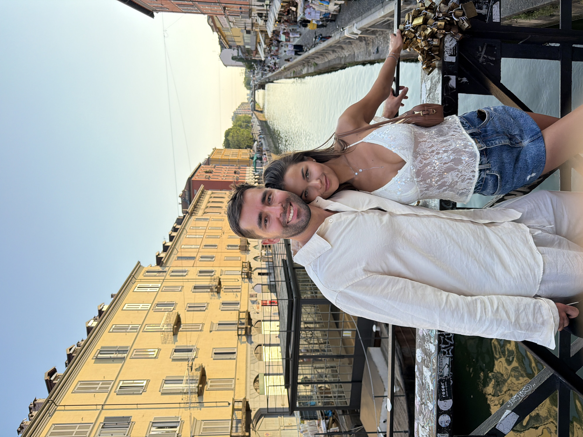 This image captures a romantic moment between a young couple on a bridge overlooking a canal in Milan, Italy. The man, wearing a light-colored button-down shirt and shorts, smiles at the camera, while the woman, in a white sequined lace top and denim skirt, leans into him. The background features charming buildings lining the canal, their facades bathed in the warm glow of the setting sun. On the right side of the bridge, a collection of love locks adorns the railing, a common symbol of commitment. The canal itself reflects the vibrant colors of the city, creating a picturesque scene. This location is likely the Naviglio Grande, one of Milan's most famous canals, known for its lively atmosphere and picturesque charm.