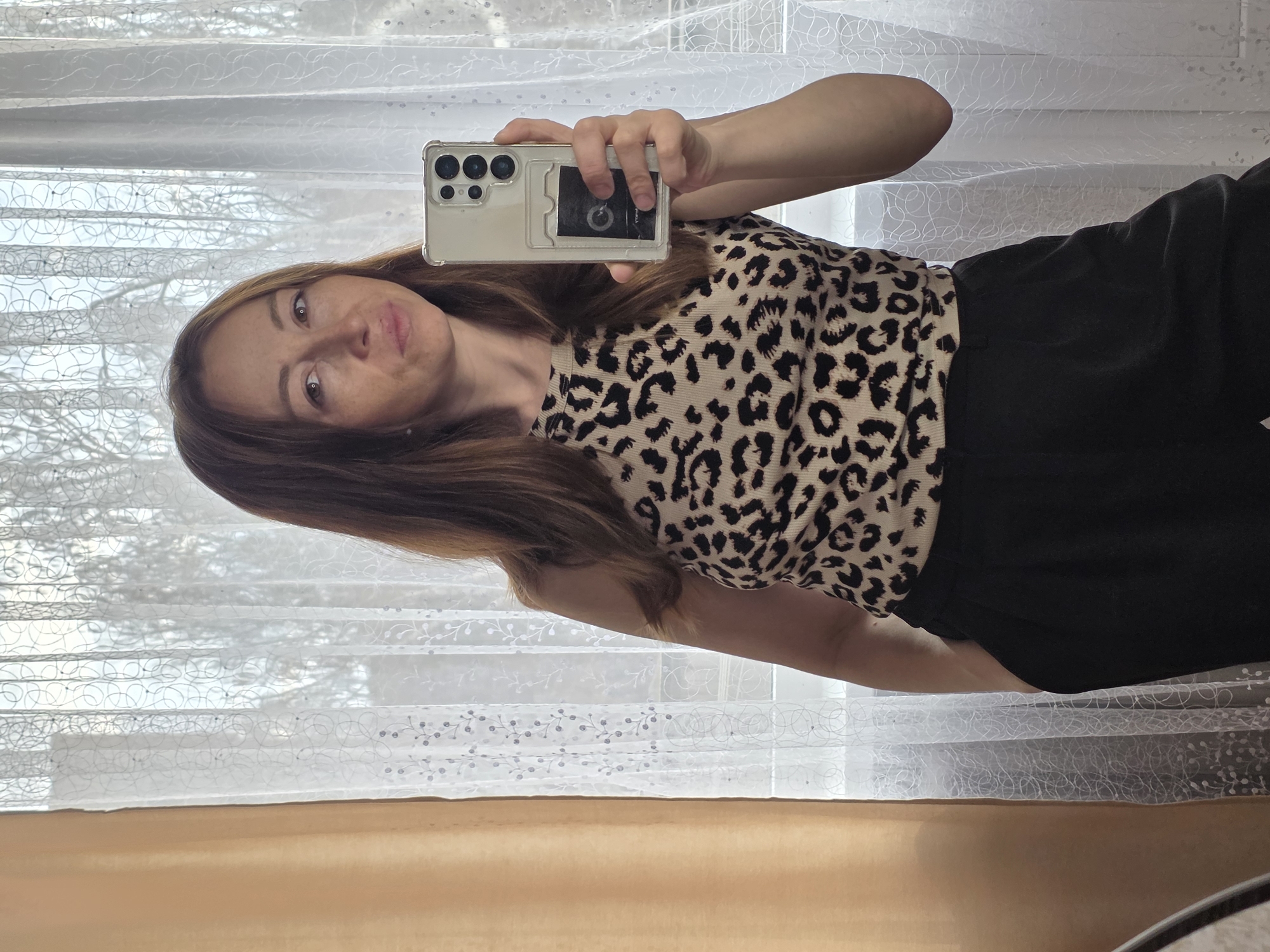 A woman is taking a mirror selfie indoors in Chaikovsky, Russia. She is positioned facing the camera, wearing a beige and black leopard print top and black bottoms. Her long, reddish-brown hair falls over her shoulders. She holds a light-colored smartphone, possibly a Samsung Ultra series, in a clear case with a card holder on the back, up to capture her reflection. Her expression is neutral, and she looks directly into the phone's lens.
Behind her, a large window is visible, covered by white sheer curtains featuring a delicate pattern of interlocking circles and floral designs. Through the curtains, the muted light of an overcast daytime is visible, along with the faint outlines of bare tree branches, suggesting winter or early spring. Below the window, a light brown or beige surface, possibly a window sill or part of the wall, extends across the bottom of the frame. No text is visible in the image.