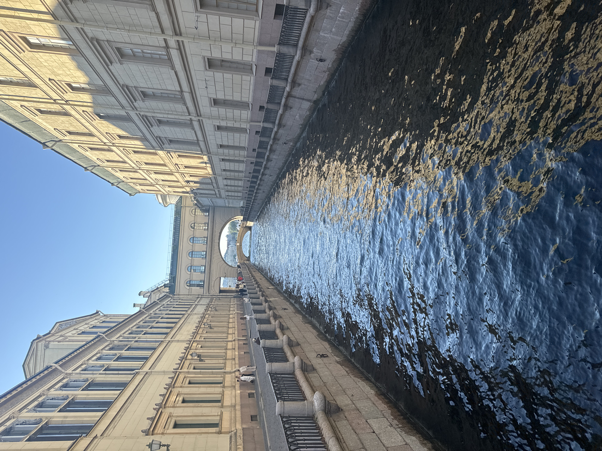 The image shows a canal in Saint Petersburg, Russia, likely the Griboyedov Canal, bordered by historic buildings. The canal water is a deep blue, reflecting the sunlight and the shadows of the surrounding architecture. Along the canal's edge, there is a stone embankment with a black metal railing. People are visible walking along the pathway next to the canal. In the background, a bridge spans the canal, and beyond that, more buildings are visible under a clear blue sky. The architecture appears classical, with detailed facades and numerous windows. The overall scene depicts a picturesque urban waterway in a historic European city.