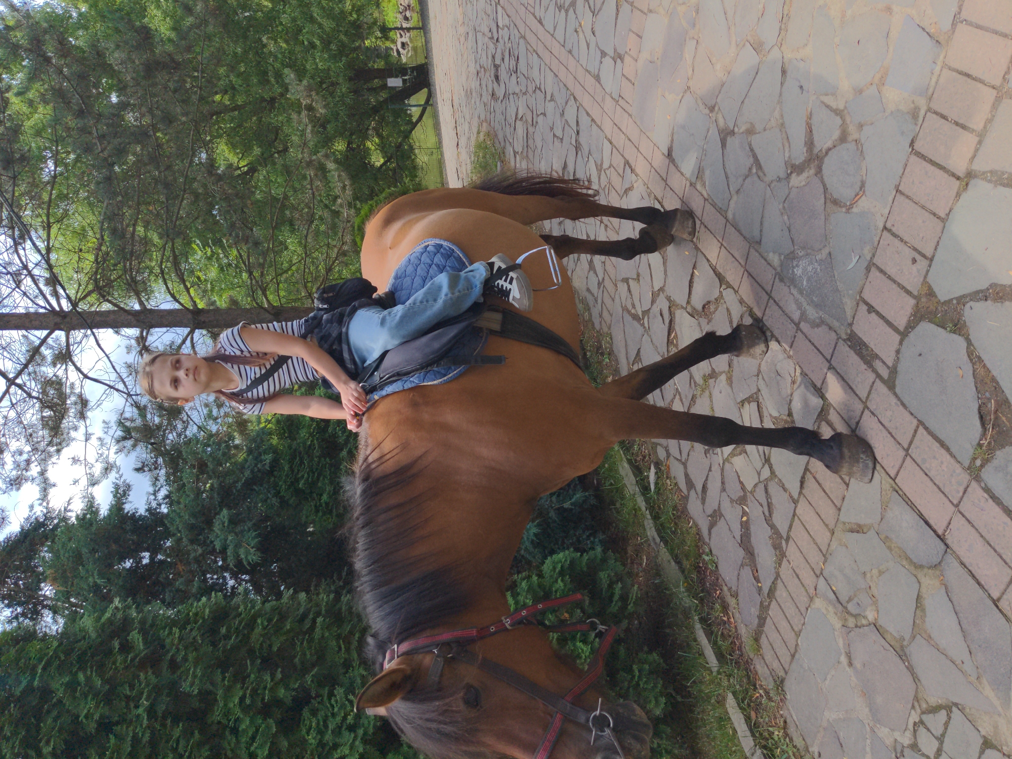 A young girl with braided hair is riding a brown horse on a paved walkway lined with trees and bushes. The girl is wearing a striped t-shirt, blue jeans, and sneakers. She has a black bag slung over her shoulder. The horse is wearing a red and brown halter. The walkway is made of irregular stone pavers and bordered by grass and low-lying shrubs. The background shows a dense foliage of evergreen trees and some deciduous trees, suggesting a park or wooded area. A pond or body of water is visible in the distance to the right. The scene appears to be in the city of Holubyne, Ukraine.