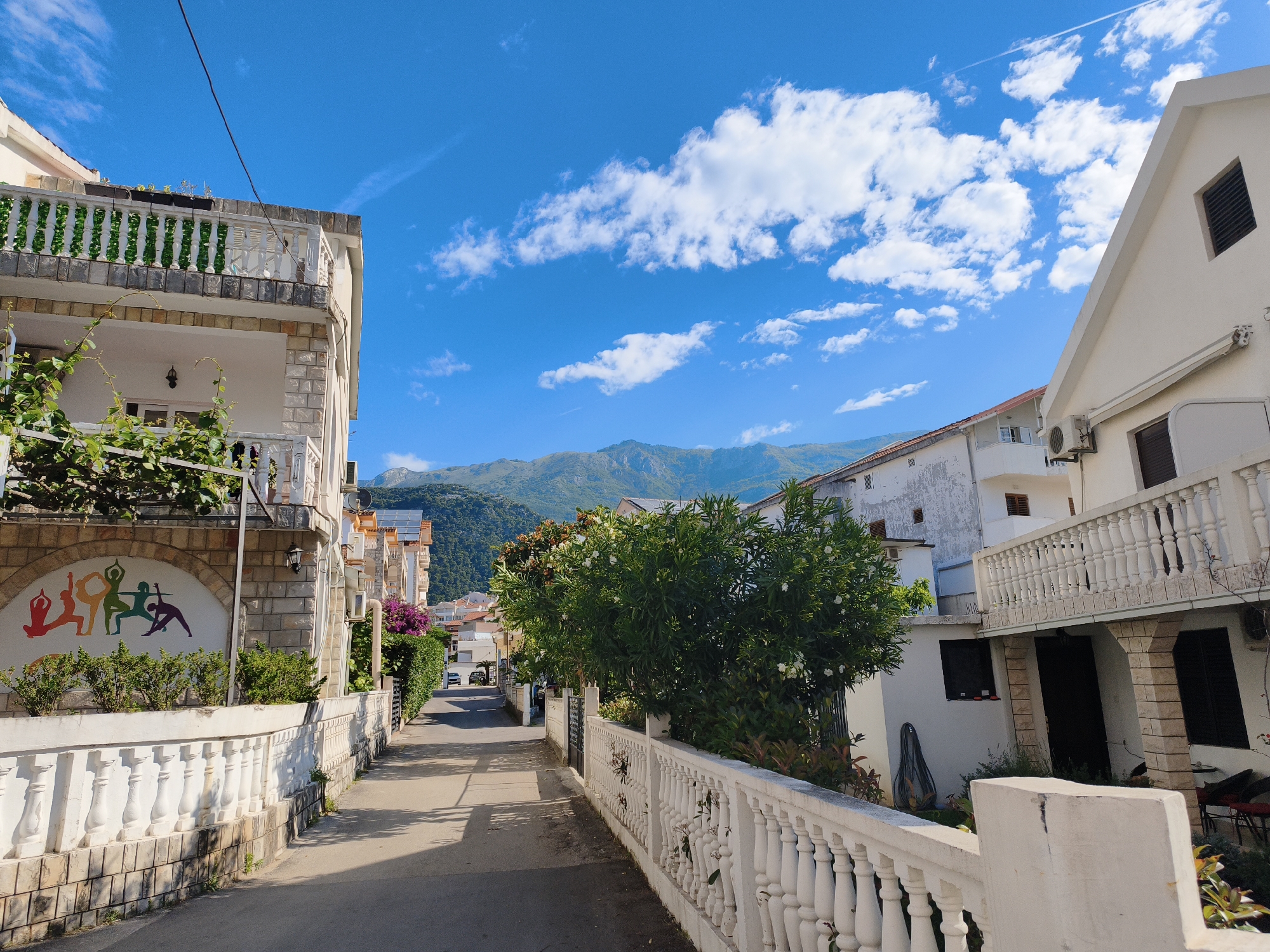The image depicts a quiet street in Budva, Montenegro. The street is flanked by buildings with white walls and white balustrade fences. A tree with white flowers sits on the right, obscuring parts of the building behind it. The street stretches into the distance, leading toward a mountain range under a blue sky with scattered clouds. On the left, a building features a colorful mural of figures in yoga poses. A utility pole is seen in the upper left corner. The street surface is concrete or asphalt. No people are visible.