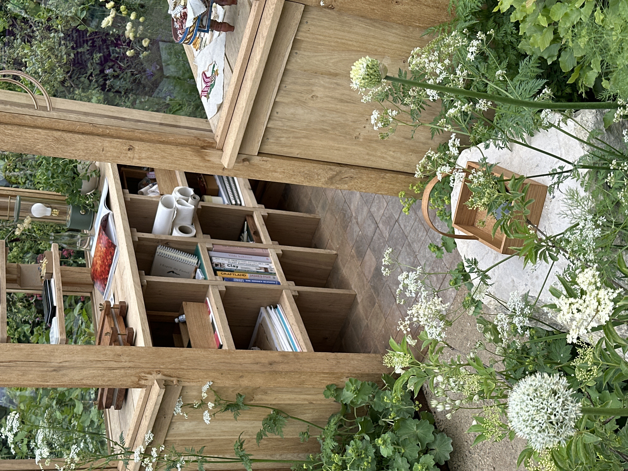 The image captures a scene that blends an interior and exterior space, likely a greenhouse or sunroom attached to a building. The main focus is on a wooden structure, seemingly a shelving unit or part of the building's framework, filled with books, rolls of paper, and other objects. The shelving unit is constructed from light-colored wood, arranged in a grid-like pattern.
Outside the wooden structure, lush greenery flourishes, with white wildflowers in bloom. The flowers frame a wooden basket sitting on a light-colored stone surface.
The flooring inside the structure is composed of diagonally laid tiles in neutral tones. Through a window at the top of the frame, glimpses of more greenery can be seen, suggesting an outdoor garden area.
While the presence of the greenhouse-like structure and the domestic objects inside indicate human activity, no people are directly visible in the frame. Given the composition of the image, and the details mentioned, it appears to be a serene outdoor space, such as a garden or patio. This image does not appear to be in London, United Kingdom.