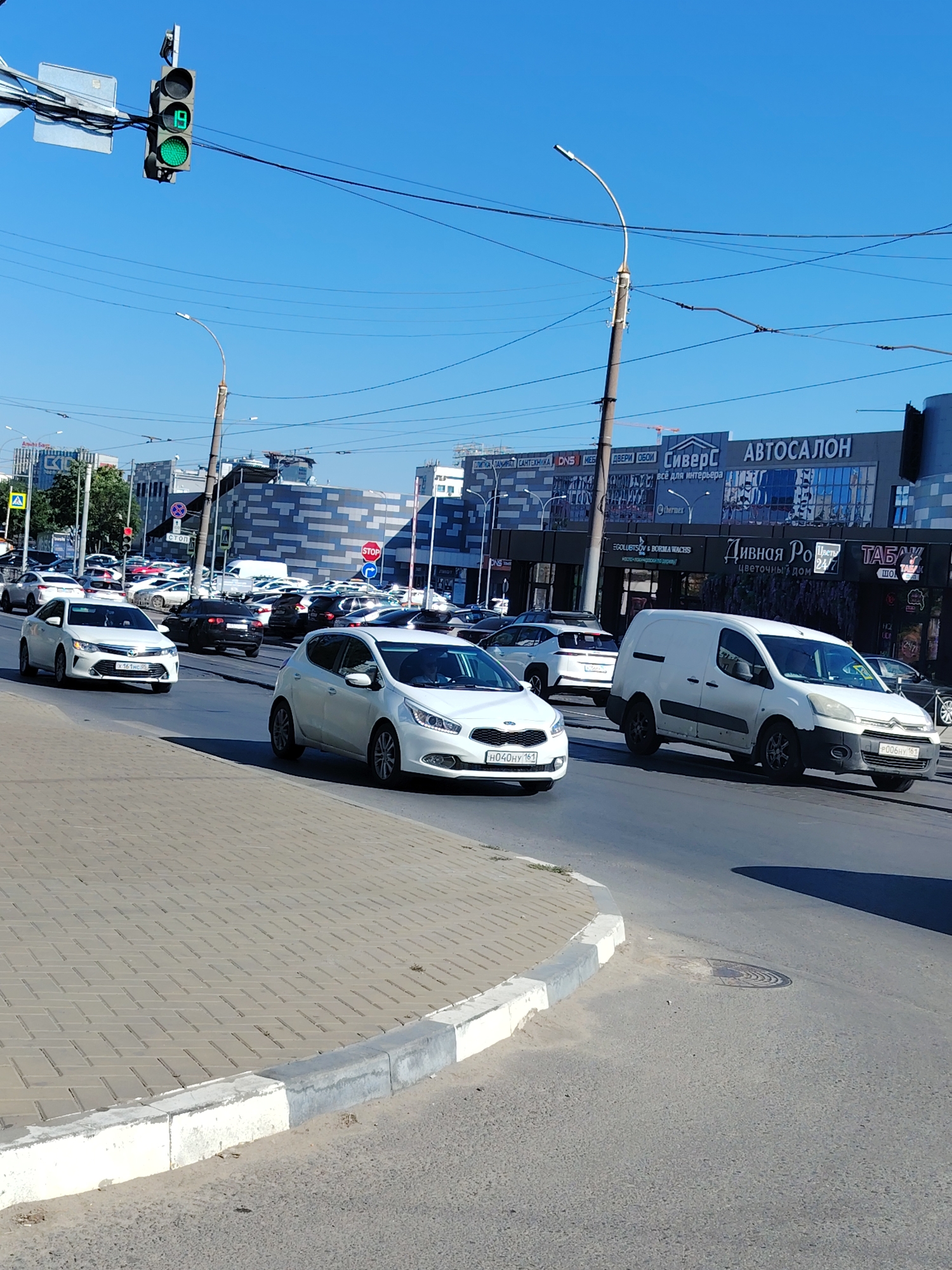 The image depicts a street scene in Rostov-on-Don, Russia, on a clear, sunny day. The foreground features a paved sidewalk with a curb bordering a multi-lane road. Several vehicles are on the road, including a white Kia hatchback, a white sedan, a black sedan, and a white van. In the background, buildings with signage are visible, including "АВТОСАЛОН" (Car Dealership) and "СиверС" (Sivers). A traffic light with a green signal is prominent in the upper left quadrant. The environment appears to be urban, with overhead wires and streetlights also visible. The traffic appears to be moving.