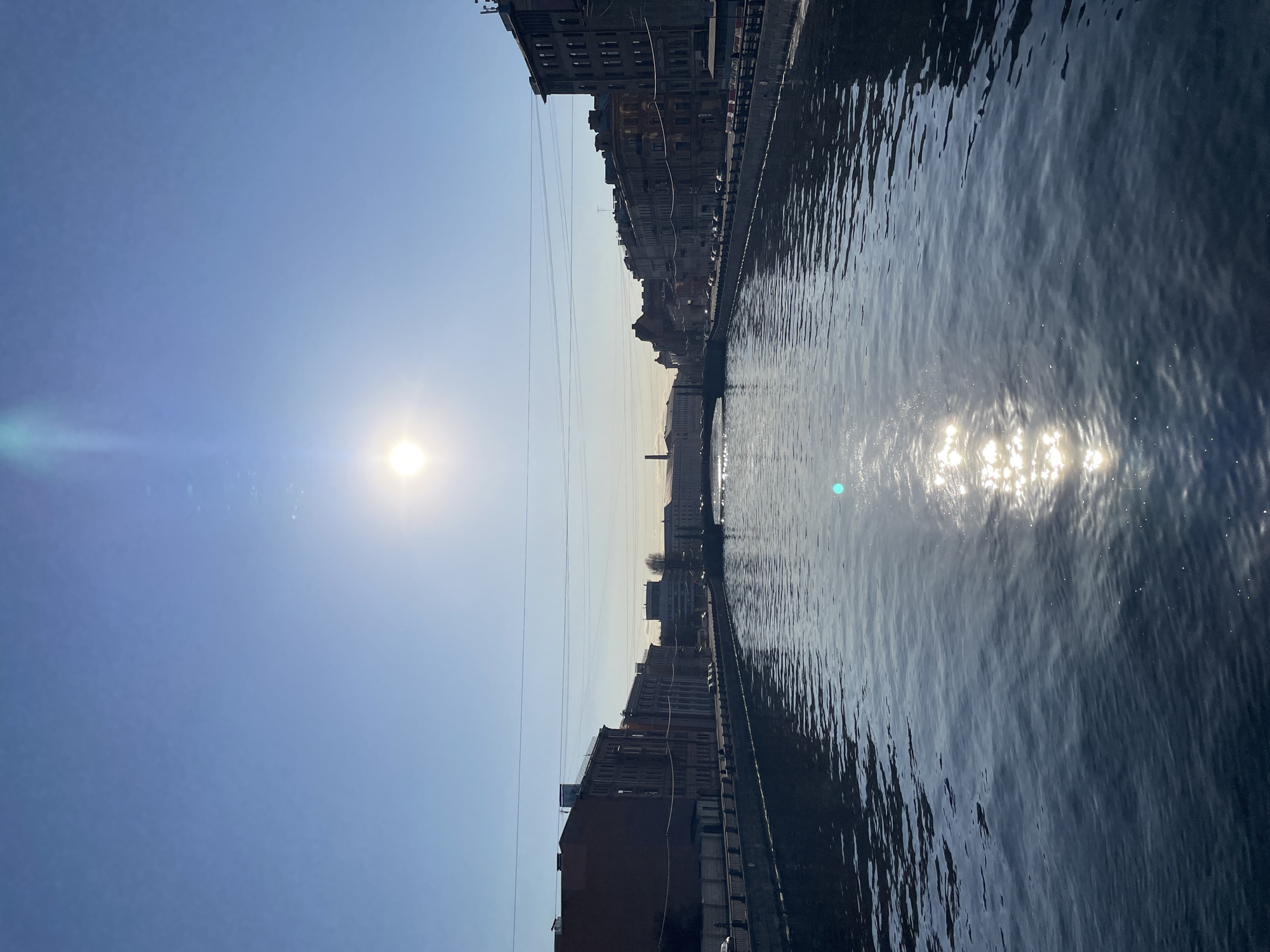 This image depicts a sunlit urban canal scene in Saint Petersburg, Russia. The perspective is looking lengthwise down a relatively straight stretch of the canal, framed by multi-story buildings on both banks.
The sky is clear and light blue, occupying the upper third of the frame. A bright, circular sun is prominently visible in the upper center-right, emitting a noticeable lens flare that extends downwards and slightly to the left.
The canal water, dark blue-grey in color, occupies the majority of the lower two-thirds of the image. Its surface is rippled, and a strong, elongated specular reflection of the sun creates a shimmering path extending from the upper-center towards the foreground.
Both sides of the canal are lined with stone or concrete embankments. Along the left bank, a row of multi-story urban buildings with historical architectural styles is visible, presenting facades in reddish-brown and gray tones, partially illuminated by the direct sunlight. A street or paved area runs between these buildings and the canal, with several parked passenger vehicles visible. Overhead power lines span horizontally across the canal at mid-height, appearing to originate from or run parallel to the left bank.
Along the right bank, a similar sequence of multi-story urban buildings is present. These buildings appear darker, being largely in shadow due to the sun's position. A street with parked vehicles is also visible along this side.
In the mid-distance, an arched bridge spans the canal, connecting the two banks. Beyond this bridge, more distant urban structures and buildings are visible, receding into a slight atmospheric haze. No individuals or active movement (e.g., moving vehicles, pedestrians) are discernible within the frame. The scene is static under clear, sunny conditions.