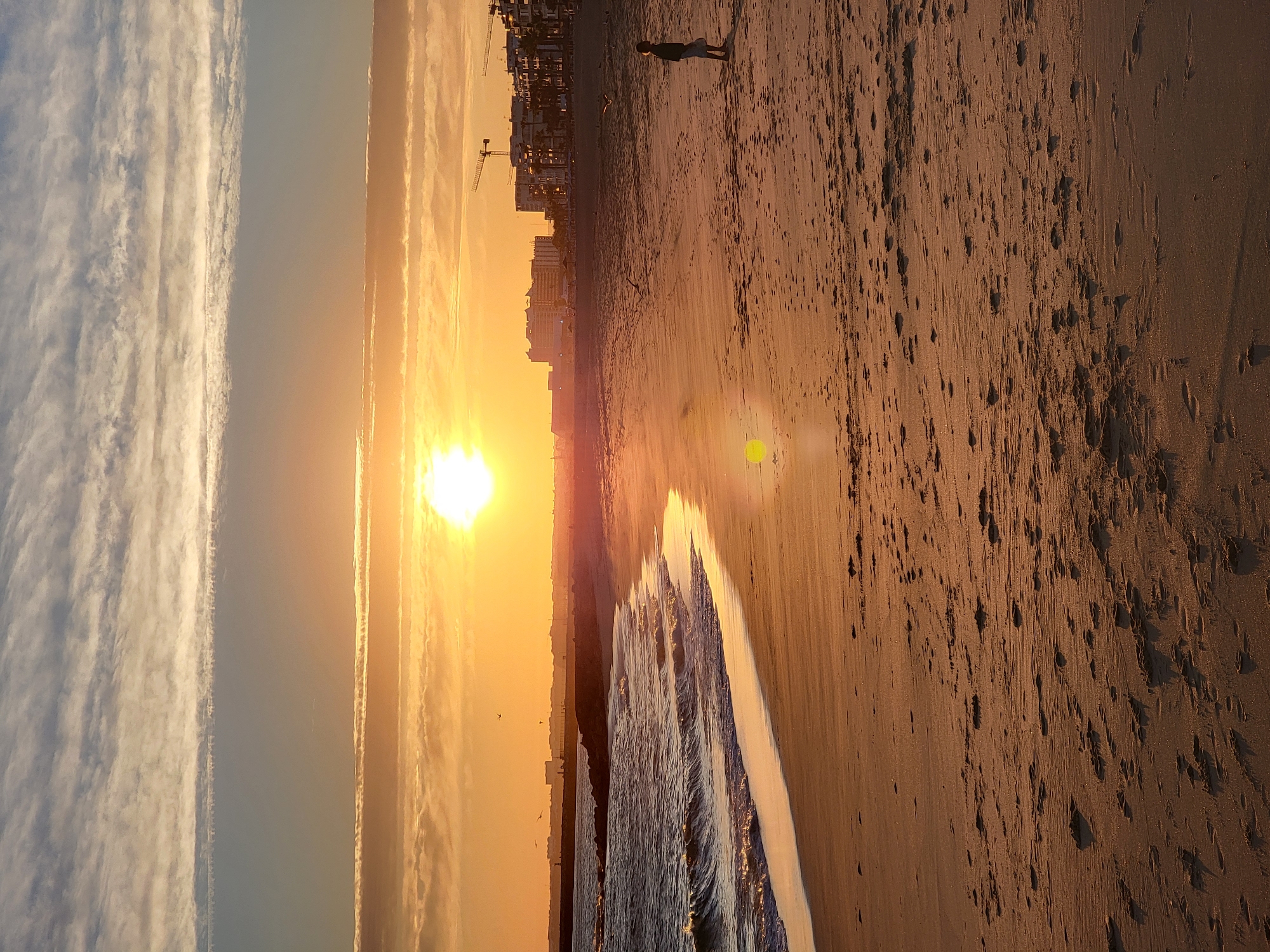 The image captures a tranquil sunset scene on the beach in Quarteira, Portugal. The sun, a bright orb of golden light, is low on the horizon, casting a warm glow across the sky and the sandy shore. Wispy clouds stretch across the sky, tinged with shades of orange and yellow from the setting sun.
In the foreground, the gentle waves of the Atlantic Ocean lap at the wet sand, creating a shimmering effect. Footprints are visible in the sand, suggesting recent activity on the beach. A lone child stands on the beach, silhouetted against the bright sky, looking out towards the sea.
In the distance, a breakwater extends into the ocean, and beyond that, the cityscape of Quarteira is visible. Buildings of various heights, including some under construction with cranes, line the waterfront. Seagulls can be seen flying in the sky. The overall atmosphere is one of peacefulness and natural beauty, characteristic of a sunset on the Algarve coast.