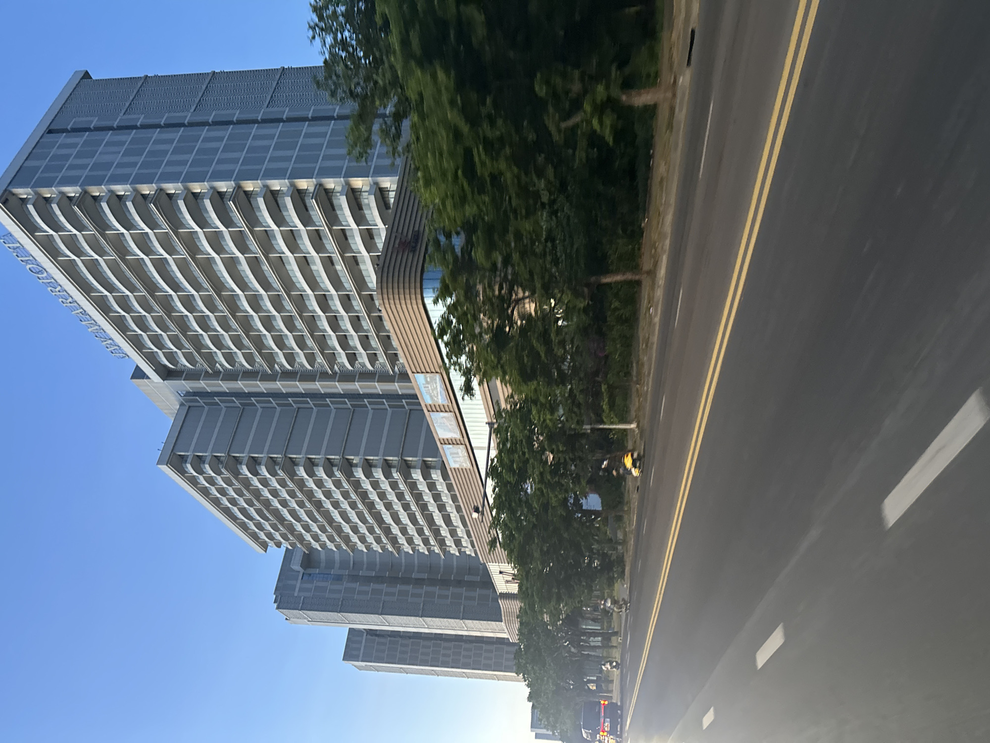 The image shows a view of the "The Filmore Da Nang" hotel, located at 16 Trần Phú, Thạch Thang, Hải Châu, Đà Nẵng 550000, Vietnam. The hotel is seen from street level, and the photograph seems to have been taken from a moving vehicle, as the road is visible in the foreground. The sky is clear and blue, with the sunlight reflecting off the road. Along the street, there are trees. In the distance, there appear to be several vehicles.