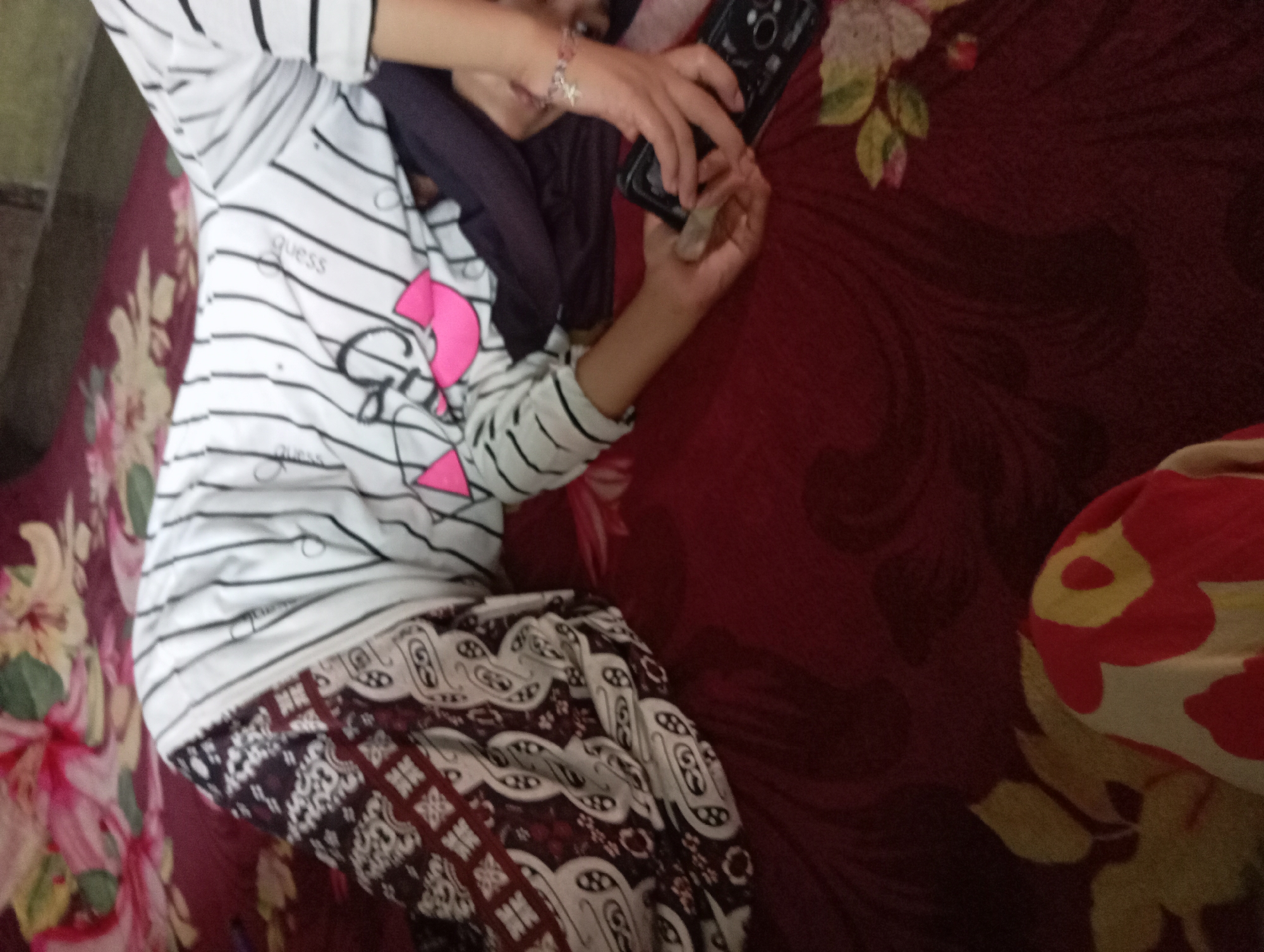 In Rejosari, Indonesia, a person is lying down on a red patterned bed. They are wearing a white t-shirt with black stripes and a pink graphic, and a batik-patterned skirt. Their right hand is holding a black smartphone, while their left hand is resting on their lap. The background consists of a floral-patterned bedspread in shades of red and pink.