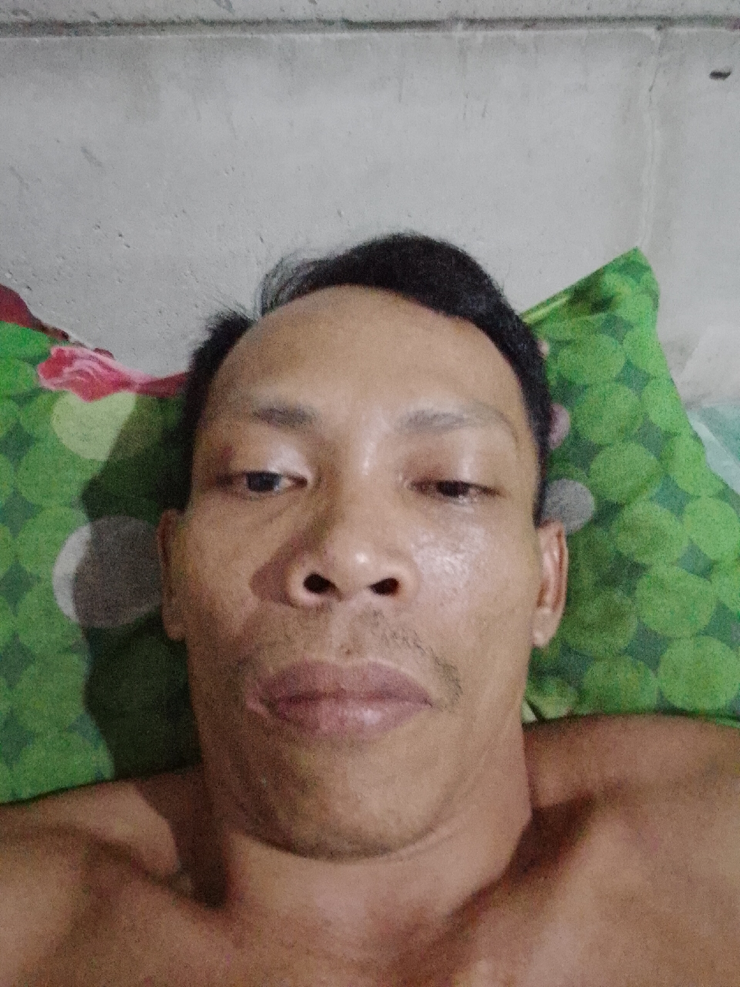 A close-up selfie of an Asian man lying on a green patterned pillow. He has dark hair, and a light complexion, and is shirtless. The background appears to be a plain, off-white wall. It is presumed the location is Bukit Ketok, Indonesia.