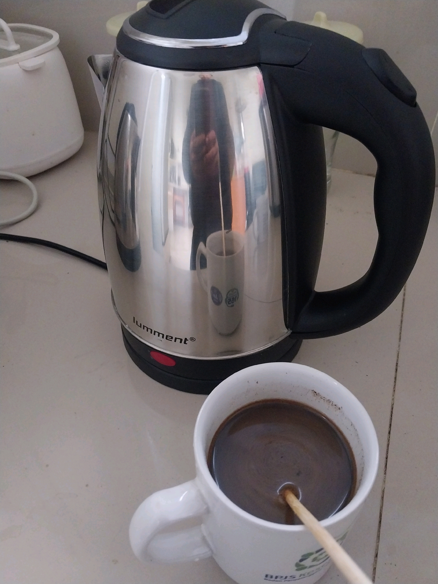 An electric kettle and a mug of coffee sit on a tiled countertop. The stainless steel kettle has a black handle and lid. A red power button is visible on the base of the kettle. The mug is white and contains dark coffee. A wooden stirrer is in the coffee. The scene is likely in a kitchen in Sleman, Indonesia, as indicated by the text on the mug.