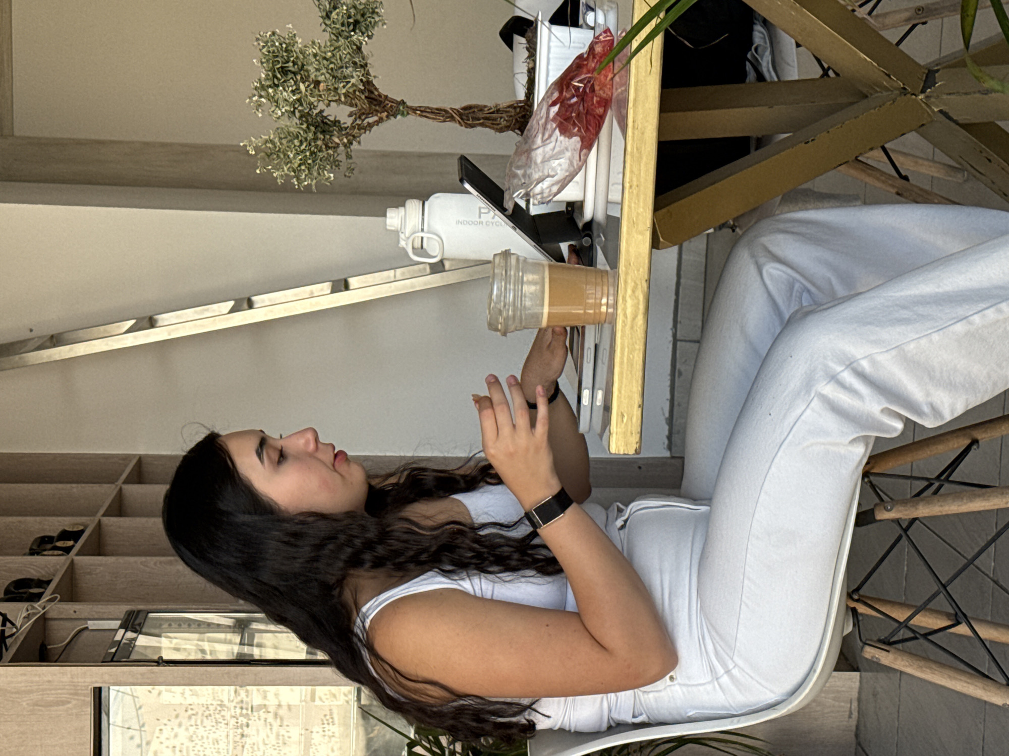 A young woman with long, dark, wavy hair is seated at a table, looking at her smartphone. She is wearing a white tank top and white pants, and she sits on a modern white chair with a distinctive wooden and black wire base. Her hands are positioned as if she is actively typing or scrolling on her phone.
On the table next to her, there is a clear plastic cup filled with a light brown beverage, likely iced coffee or tea. A white water bottle, partially obscured but showing the text "P" and "INDOOR CYCL" on its side, stands near a smartphone in a black case, which is propped up. Further back on the table, a small decorative plant, resembling a bonsai, is visible, along with a clear plastic bag containing reddish items.
The background features a light-colored wall and a wooden shelving unit with multiple open compartments, some containing footwear. A gold-colored table leg or frame is also prominent. The scene is brightly lit, suggesting it is daytime. The woman appears calm and focused on her device within a contemporary and relaxed setting.