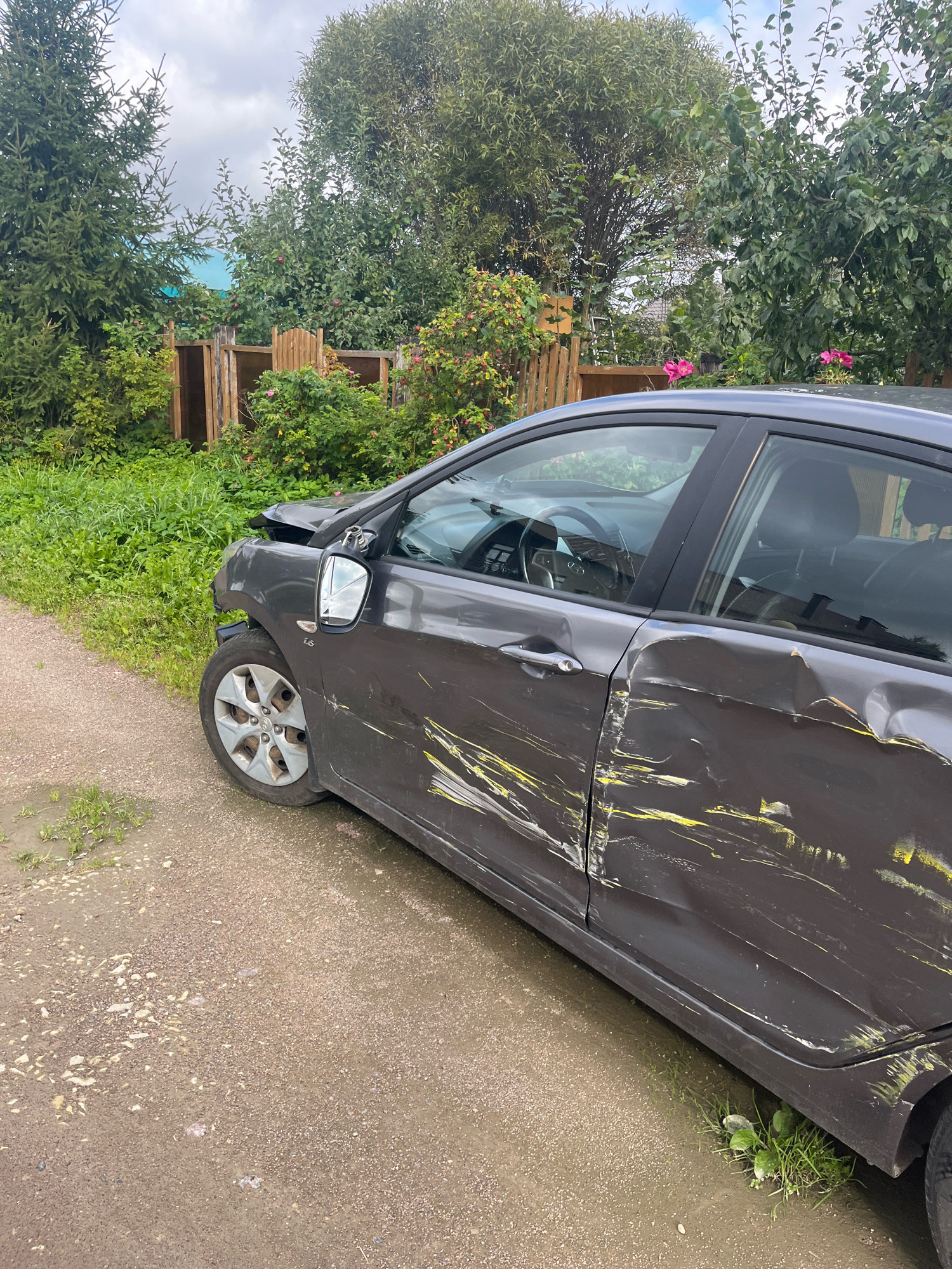 A dark gray sedan, heavily damaged on its driver's side, is parked on a gravel road adjacent to a grassy area. The front fender and hood are crumpled, and the driver's side door is significantly dented and scratched, with yellow paint marks visible. The side mirror is also damaged. The car is positioned at an angle, facing towards the right of the frame. The background consists of lush green trees, a wooden fence, and some pink flowers. The sky is overcast. The scene appears to be in a rural or semi-rural setting, possibly Vladimirovka, Russia, given the presence of typical Russian dacha-style fencing and vegetation. No people are visible in the image.
