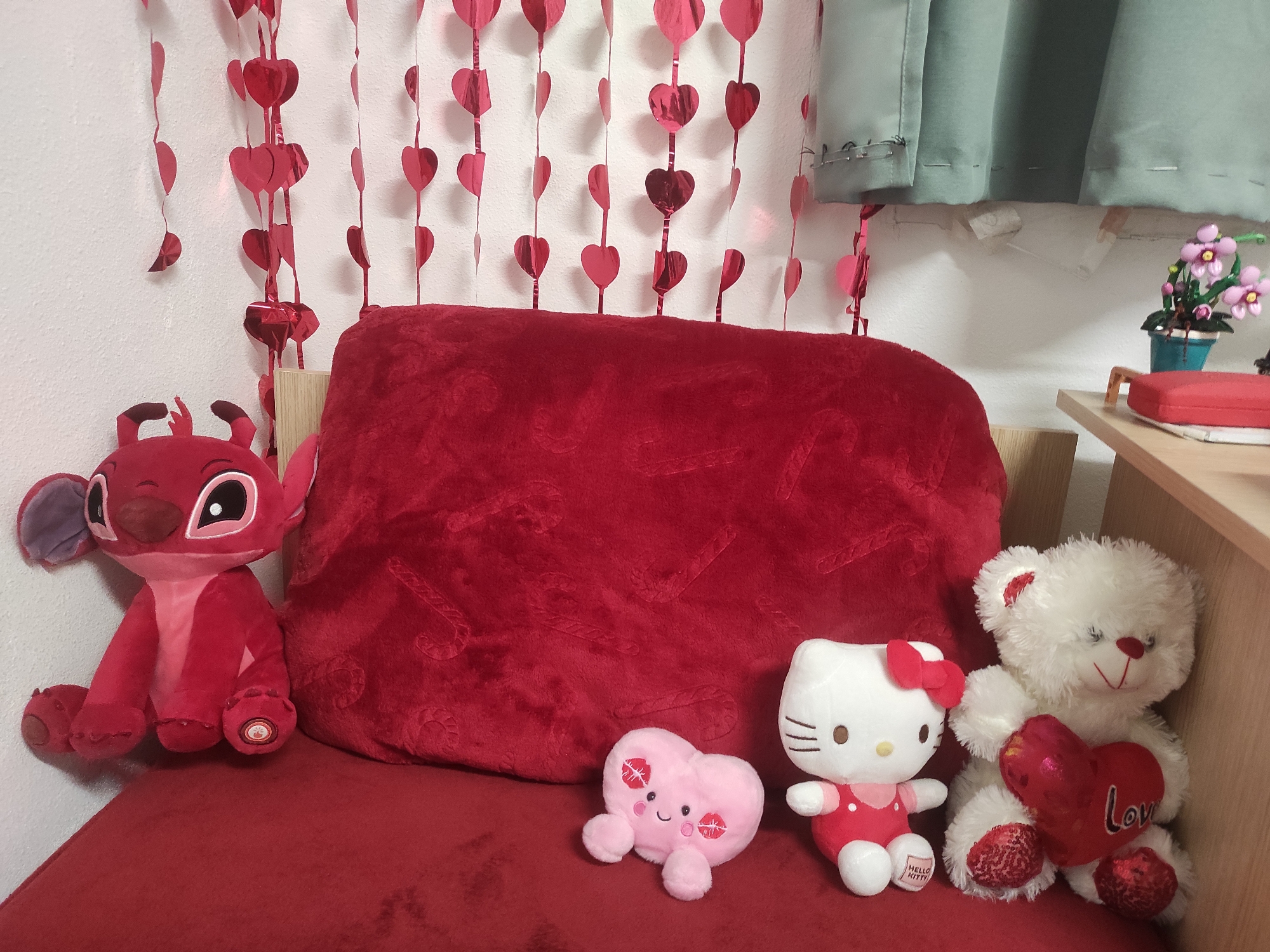 The image depicts a collection of plush toys arranged on a red cushion, likely part of a couch or seating arrangement. The toys include a red Stitch plush, a pink heart-shaped plush with kiss marks, a Hello Kitty plush in a red dress, and a white teddy bear holding a red heart with the word "Love" on it. A large red, plush pillow is placed behind the toys. The background includes hanging strings of red heart decorations, a white wall, and a glimpse of a green curtain. A small shelf with flowers and a red object are visible on the right side of the frame. The location cannot be determined from the image.