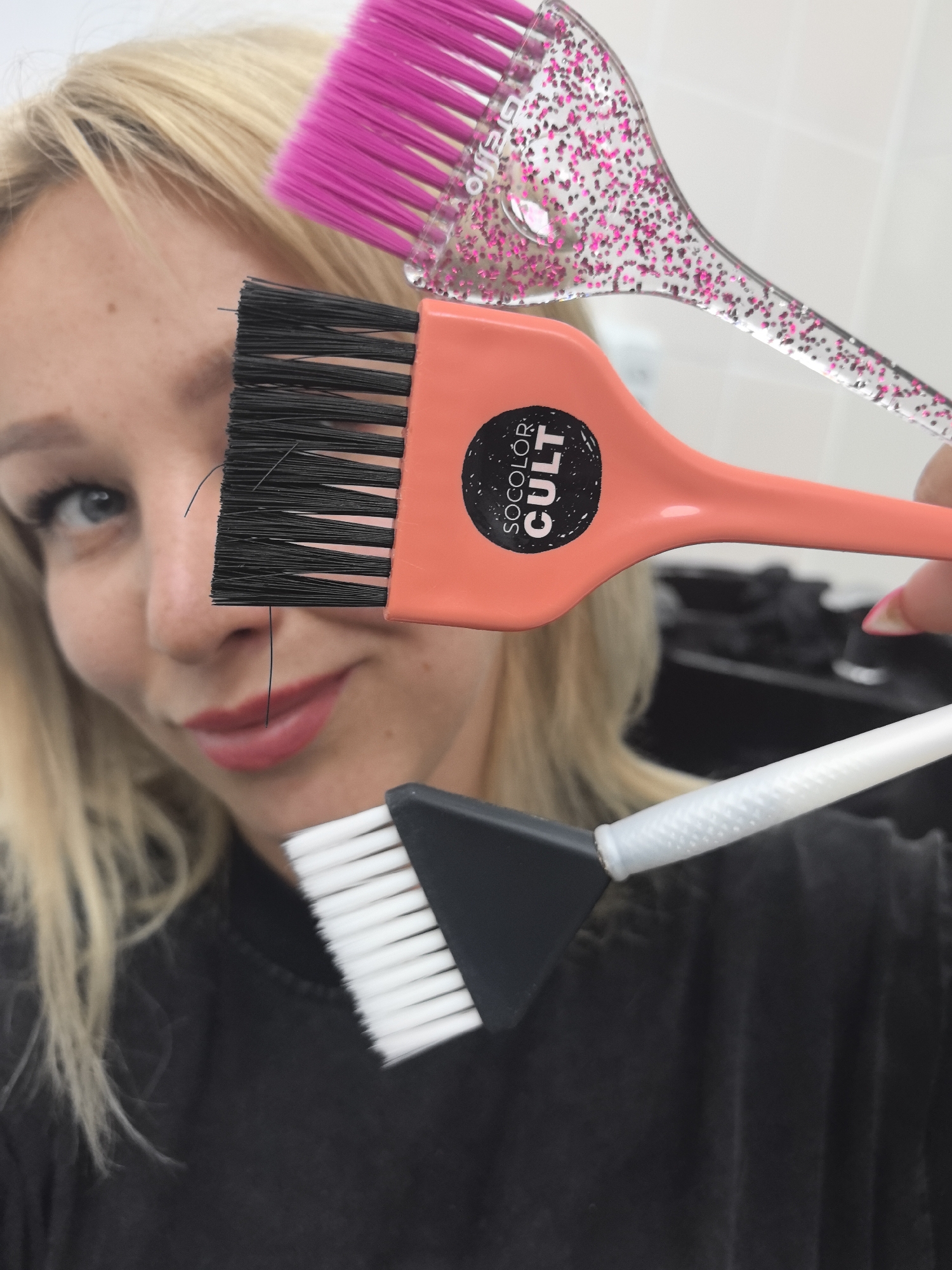 In this close-up, vertical-oriented photograph, a fair-skinned woman with blonde hair and blue eyes is partially visible. She is looking slightly off-camera, and her lips are painted with red lipstick. She is holding three hair coloring brushes. The topmost brush has a clear handle with pink glitter and pink bristles. Below it is an orange brush with a black label that reads "SOCOLOR CULT" and has black bristles. The bottom brush has a black handle and white bristles. The background is out of focus, but appears to be an indoor setting, possibly a salon, with dark-colored equipment visible behind the woman. There is no indication of the city of Chaikovsky, Russia.