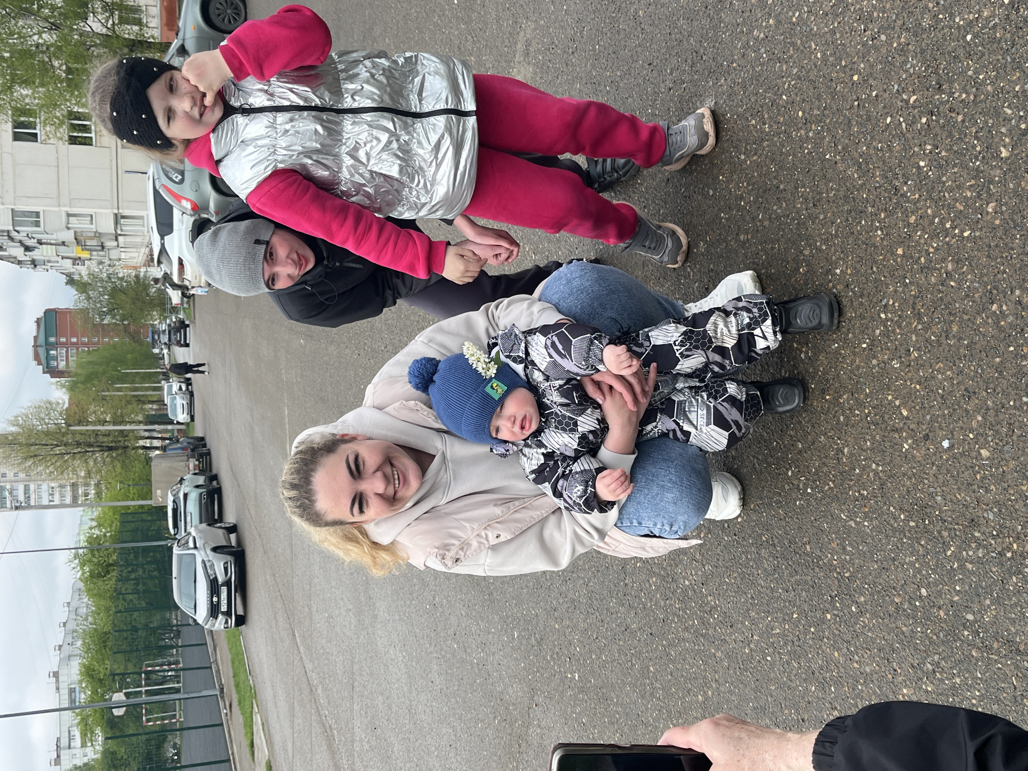 The image shows a woman sitting on the ground, holding a baby. Another woman is behind her, also on the ground, and a young girl is lying on top of the second woman. They are all smiling. The setting appears to be a street in a city. The location is identified as Chaikovsky, Russia due to the license plates visible on the parked cars. Buildings and trees are visible in the background. A person is taking a photo with a phone in the lower part of the image.