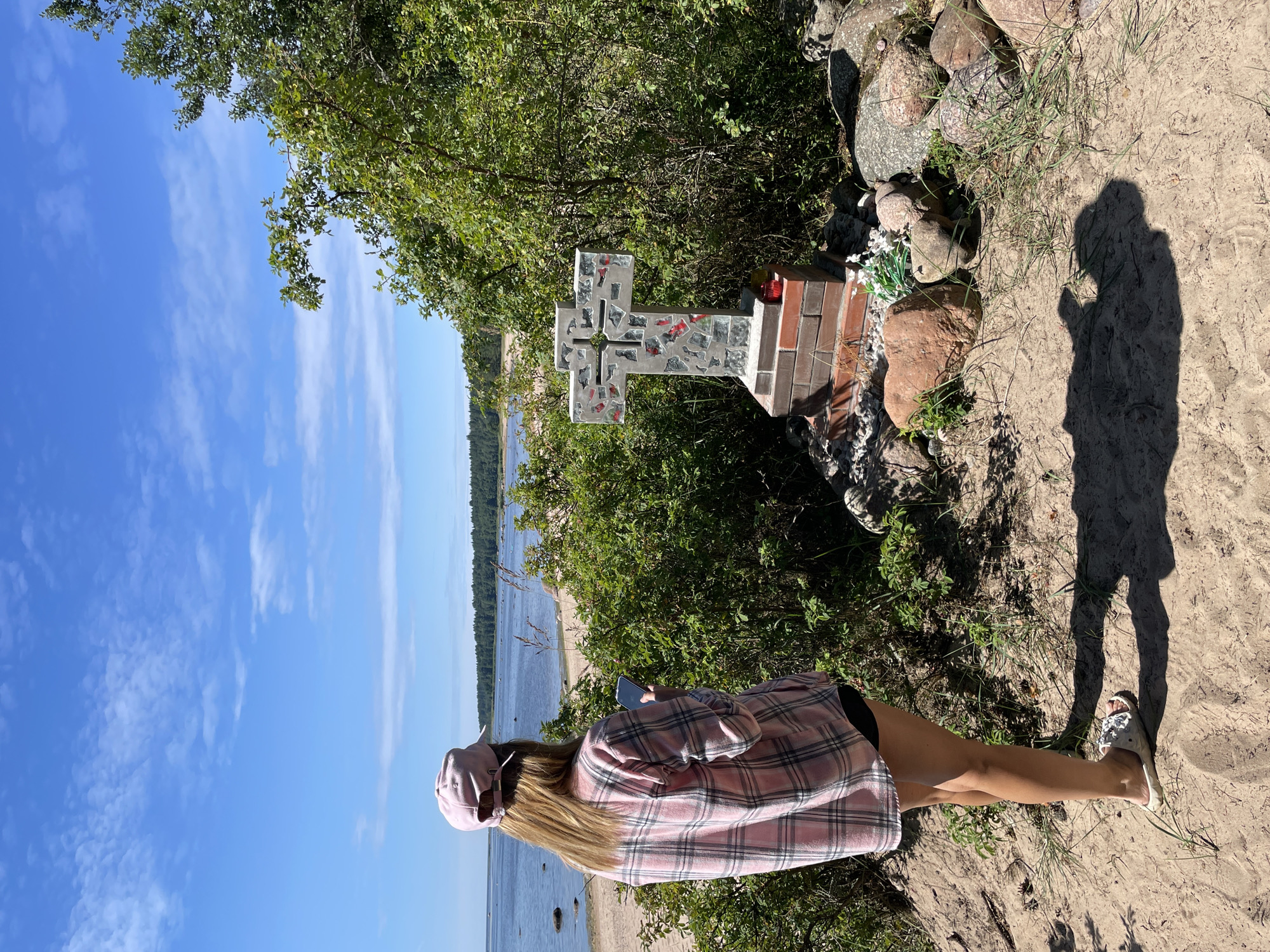 A young woman wearing a pink plaid shirt and a pink baseball cap stands on a sandy beach in Zelenaya Roshcha, Russia, holding a smartphone. She is facing away from the camera, looking out at the sea. To her right, a mosaic cross stands on a brick base, adorned with red flowers and surrounded by rocks. The sky is blue with scattered white clouds, and the sea stretches to the horizon, where a dense forest can be seen. The overall scene depicts a peaceful, natural setting.