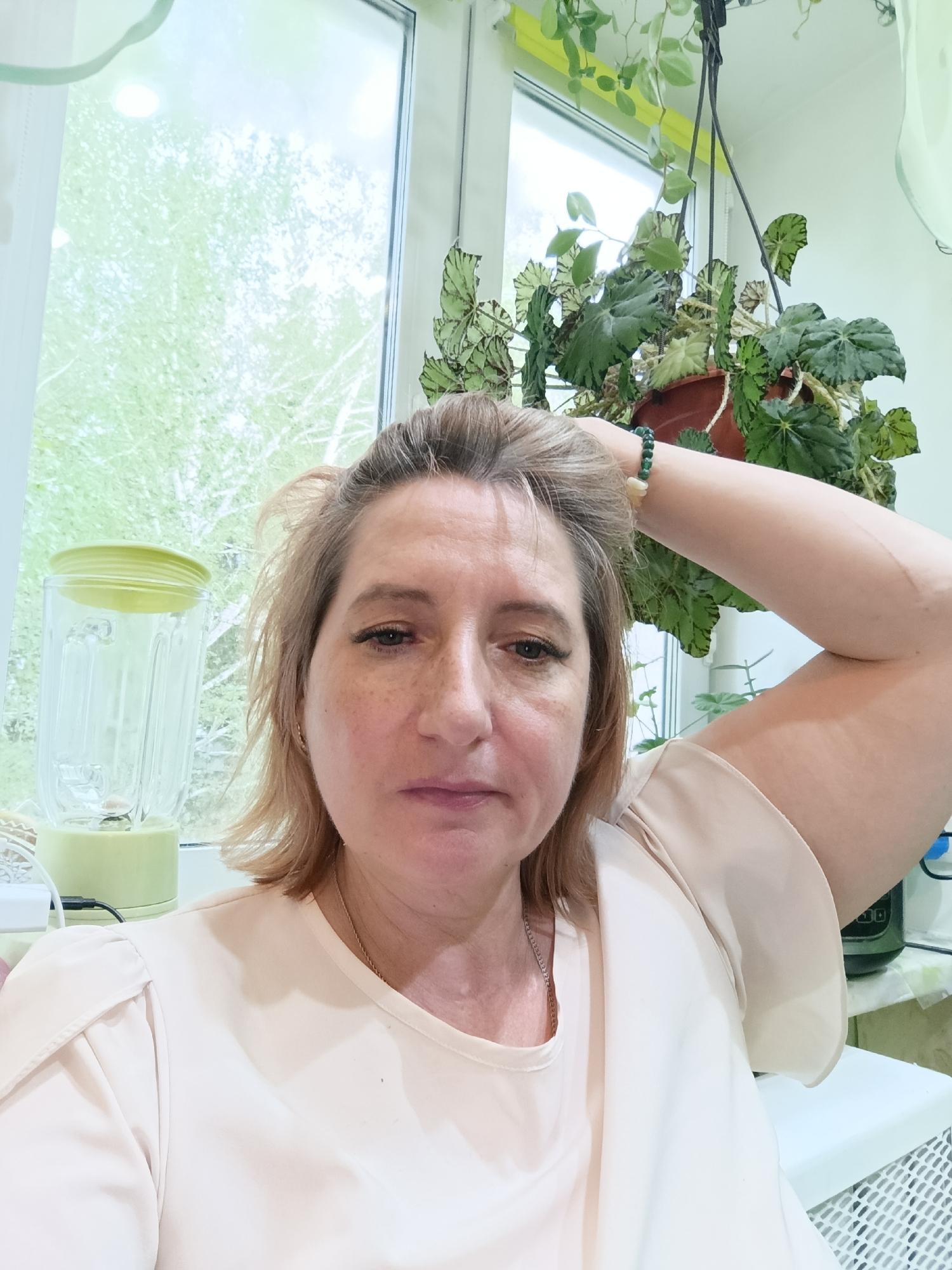 The image shows a woman with blonde hair and fair skin, posing for a selfie. She is wearing a light-colored, ruffled blouse and a silver necklace. Her right arm is raised behind her head, and she is wearing a green and yellow bracelet on her wrist. She has freckles on her face and her expression is neutral.
In the background, a window looks out onto greenery, suggesting it is daytime with good weather. To the left of the woman, a light green blender with a translucent pitcher is visible. Behind her, hanging from the ceiling, is a potted plant with large, patterned green leaves. The setting appears to be a home, possibly a kitchen or living area, with a window providing natural light.
The overall mood of the image is casual and relaxed. There is no visible text in the image. The location context provided is Tugulym, Russia.