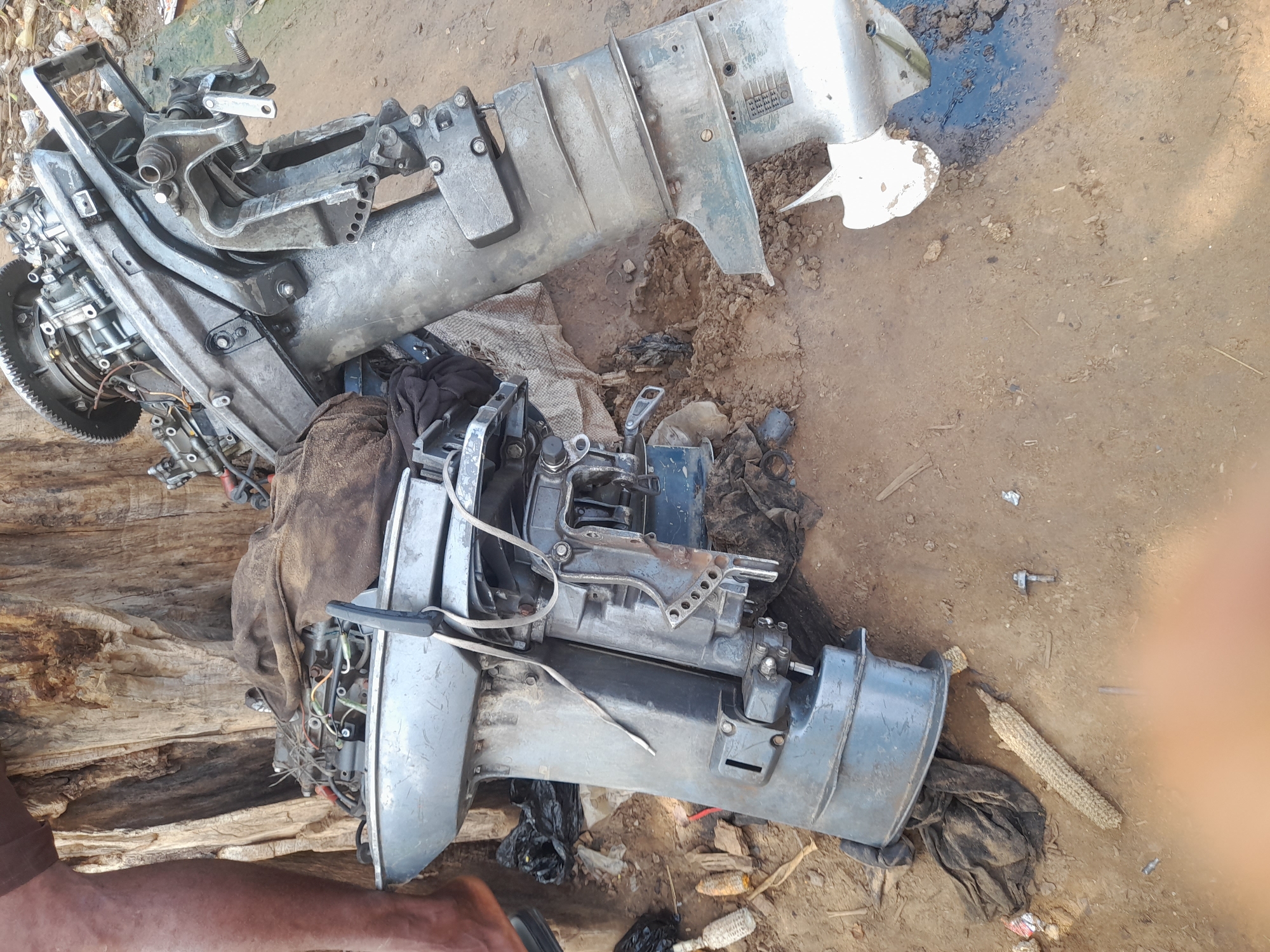 In Nemnei, Nigeria, two outboard motors are positioned on the ground. The motors are disassembled, with one motor positioned upright and the other lying on its side. One motor is gray and appears to be made of aluminum. The other motor is also gray but is darker in color and appears to be made of steel. Both motors are covered in dirt and debris. The area surrounding the motors is a dirt path, with some debris scattered around. A tree trunk is visible in the background on the left side of the image. A person's leg is visible on the left side of the image.
