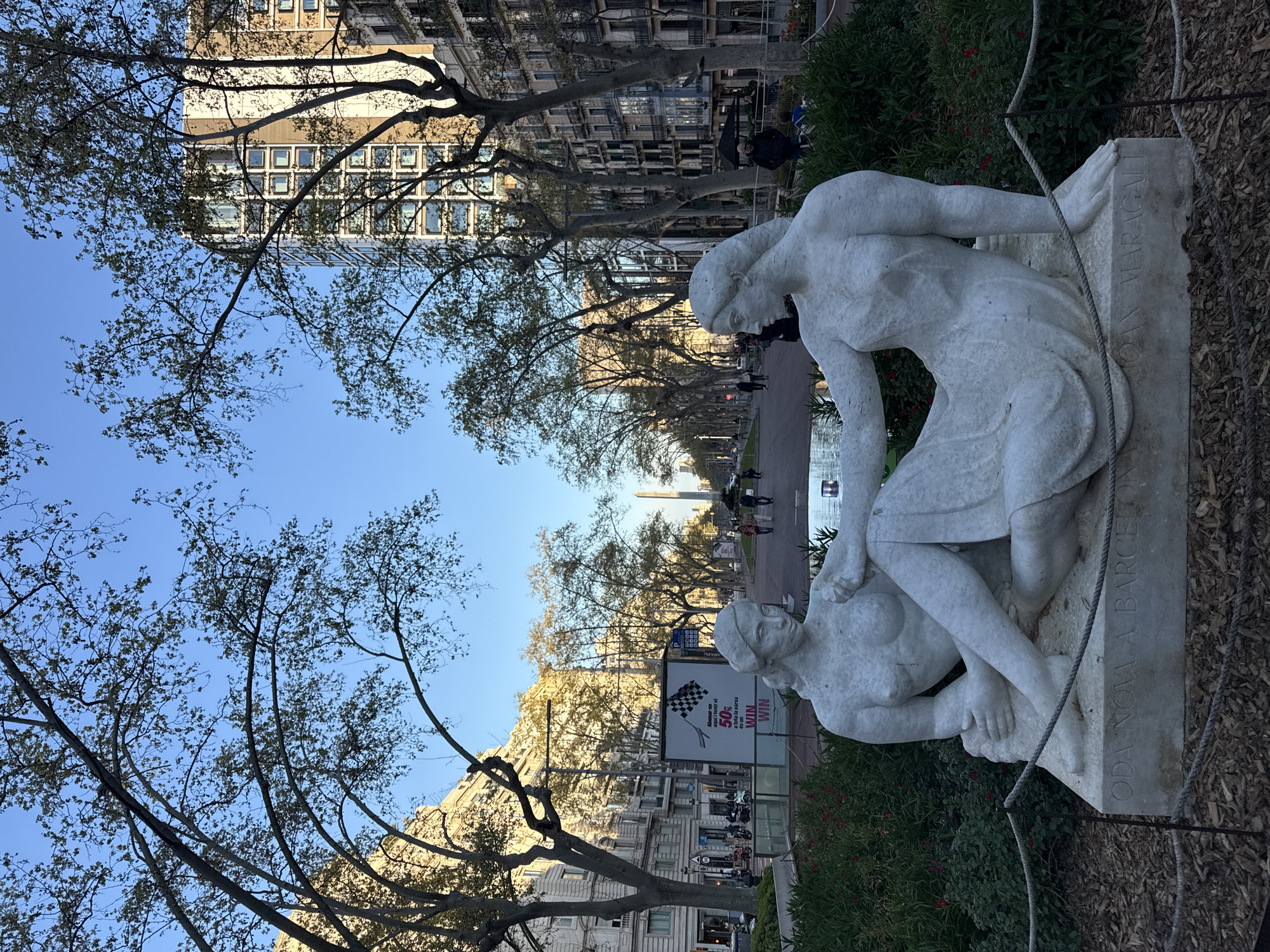 Here's a detailed description of the image:
The image captures a scene in Barcelona, Spain, centered around the "Oda Nova a Barcelona" sculpture by Joan Maragall.
Foreground:
* A white marble sculpture depicting two figures in an intimate, possibly nurturing pose dominates the foreground. The sculpture is mounted on a stone base with engraved text.
* Foreground objects in front of the statue include small rope fencing and wood chips
* Green shrubbery with small red flowers surrounds the base of the sculpture.
Background:
* The backdrop is a wide avenue featuring a tree-lined pedestrian walkway.
* Several people are walking along the avenue, some visible near a water feature and a tall, monument-like structure in the distance.
* Buildings with multi-story facades line the avenue.
* There's a large advertising poster with a checkered flag and text, indicating a promotion related to subscription services.
* The sky is clear and blue, visible through the branches of the trees lining the avenue.