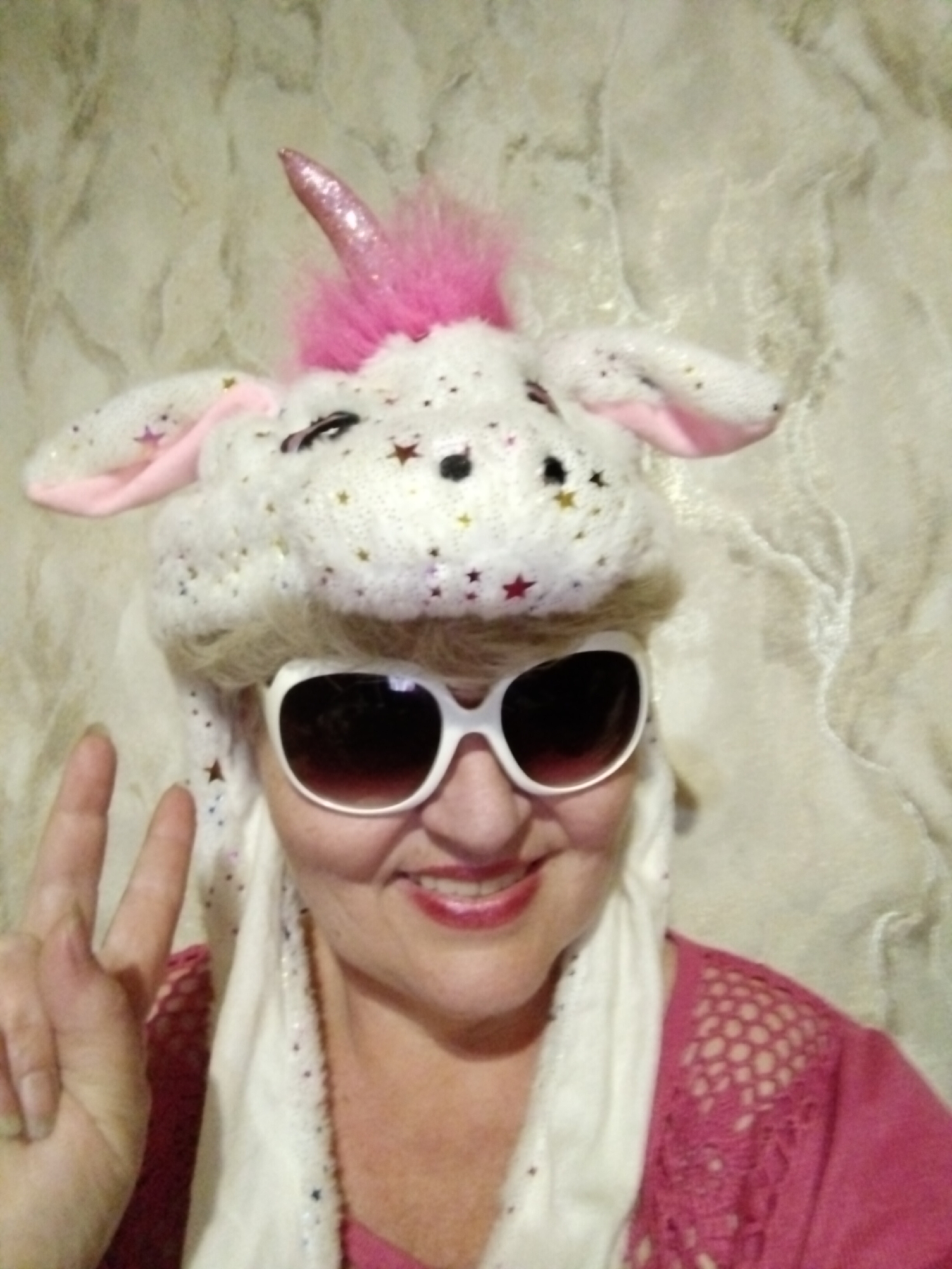A person is wearing a white and pink unicorn hat and white sunglasses, making a peace sign with their hand. The person is wearing a pink shirt and a white scarf. The background is a textured, light-colored wall. The location is Horishni Plavni, Ukraine.