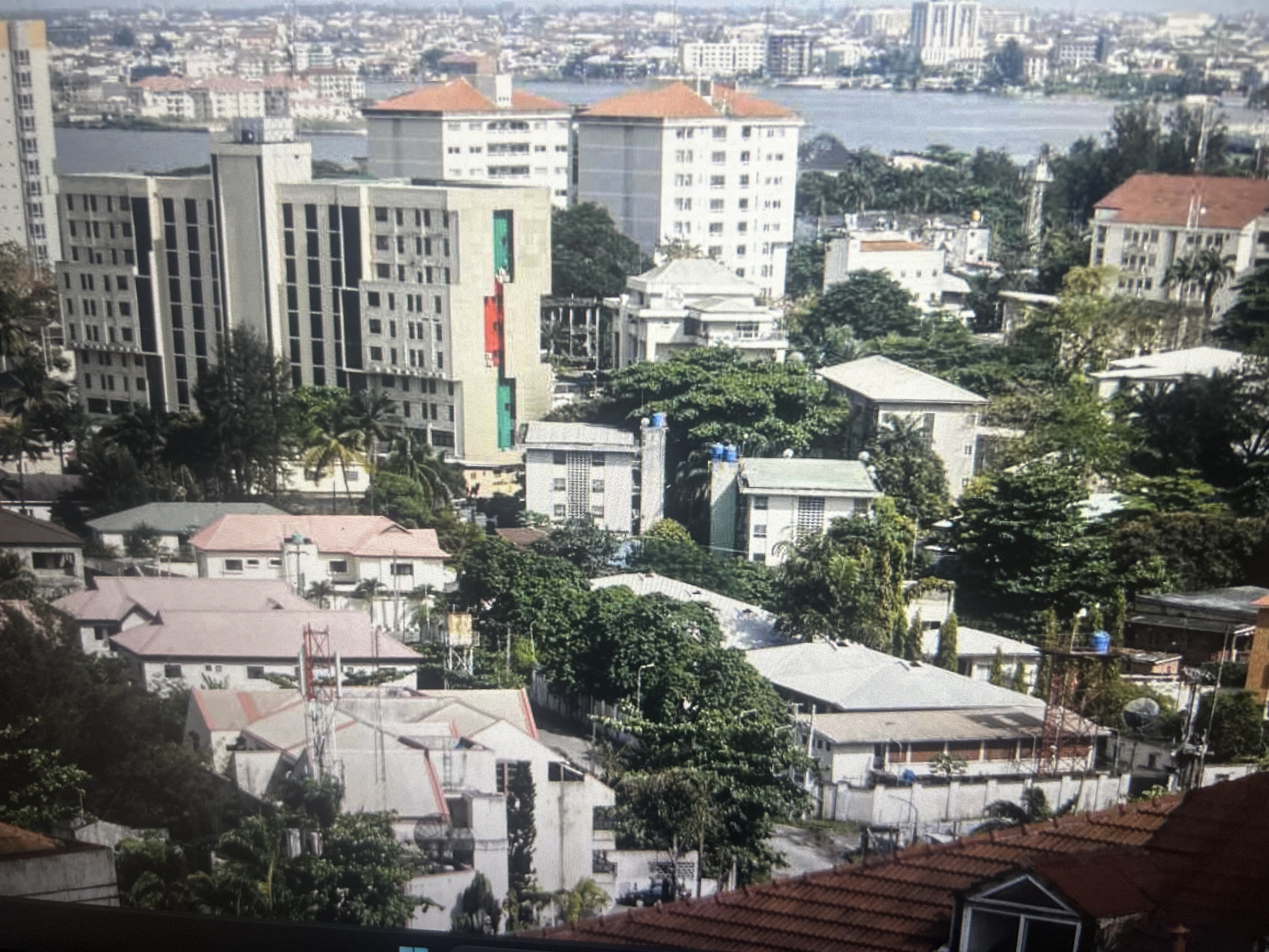 This is an aerial, full-color, slightly blurry photograph depicting a dense urban environment in Ogbomosho, Nigeria. The image is dominated by numerous buildings of varying sizes and architectural styles, with a notable presence of multi-story structures and lower-lying residential buildings. Lush green trees and foliage are interspersed throughout the scene, indicating a tropical or subtropical climate. A body of water is visible in the background, suggesting proximity to a river or lagoon. The sky is overcast, casting a diffused light on the landscape. No individuals are clearly discernible in the frame. The scene is characterized by a high density of man-made structures and abundant vegetation.