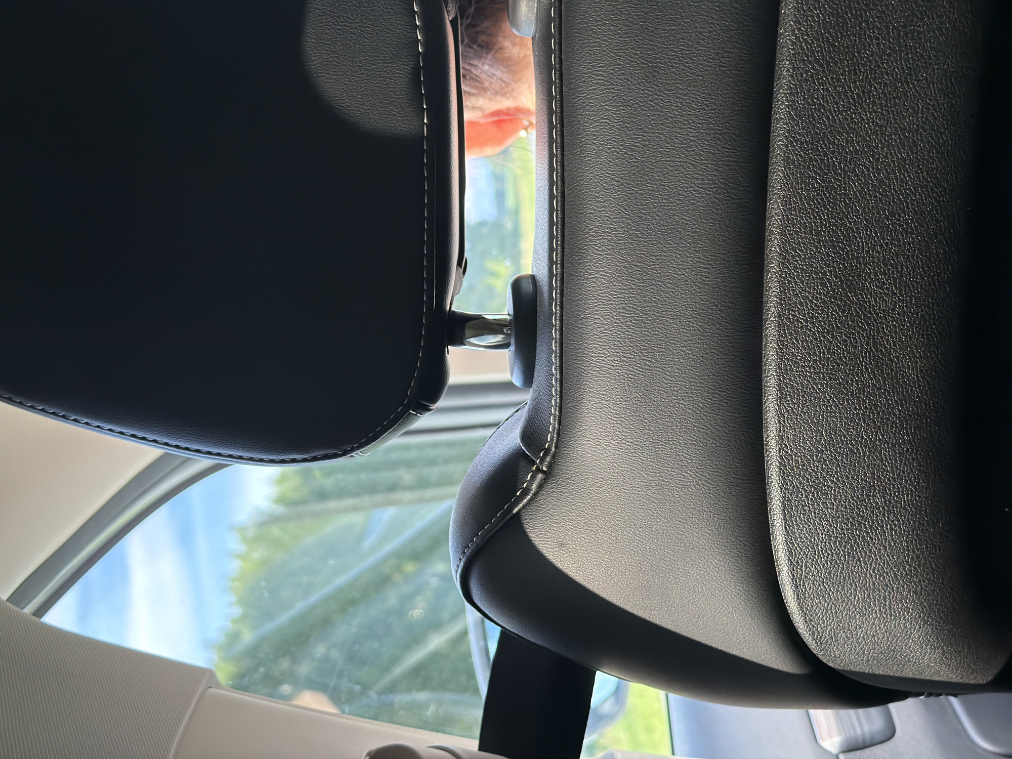 The image provides an interior view from a car, seemingly captured from a rear seat looking forward. Two dark-colored, textured headrests, likely made of leather or faux leather with distinct white stitching, are prominent in the foreground. They frame a narrow vertical gap in the center of the image.

Through this gap, a portion of a person's head is visible, specifically an ear, part of the cheek, and some light-colored hair, suggesting someone seated in front. The metallic support rods connecting the headrests to the seatbacks are also seen within this space.

Beyond the interior elements, a vibrant outdoor scene is revealed through the car window. A clear blue sky with a few wisps of white cloud is visible, along with abundant green foliage, indicating trees or bushes. The bright daylight creates strong contrasts and shadows within the car's interior. Parts of the car's light-colored interior trim and a seatbelt strap are also visible towards the bottom and left edges of the frame. The scene depicts a sunny day, with the car likely in motion or parked outdoors in a natural setting. No text is visible.