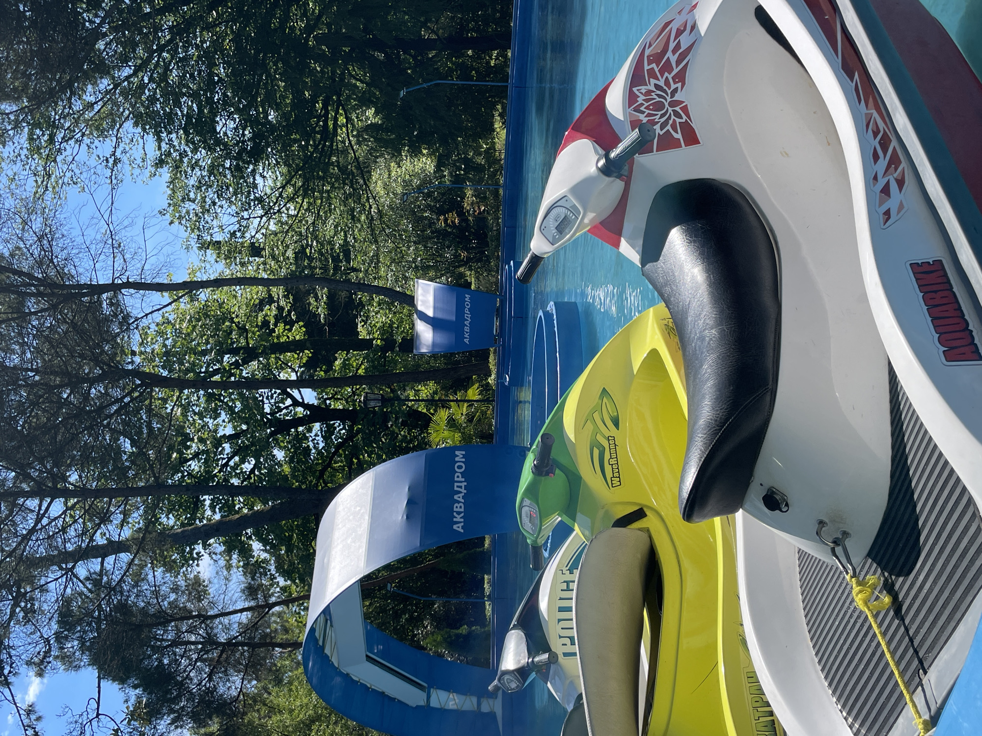 The image captures an outdoor recreational scene featuring several personal watercraft (jet skis or aquabikes) in a blue water channel, under a clear blue sky. The photo is rotated 90 degrees clockwise from its correct orientation.
In the right foreground, a white and red aquabike with a black seat is prominently displayed, bearing the text "AQUABIKE" on its side and a decorative lotus-like motif. Behind it, a yellow and green watercraft, labeled "WaveRunner," is partially visible. The watercraft are positioned in a narrow, man-made blue water channel or pool, with reflections indicating bright sunlight.
In the midground, a distinctive blue and white archway spans over the water channel. The Cyrillic text "АКВАДРОМ," which translates to "AQUADROME," is clearly visible on the archway, identifying the location as a water sports venue. Another smaller blue sign with the same text is visible further back in the channel.
The left side and background of the image are dominated by lush, dense green trees, suggesting a park-like or natural setting. Patches of clear blue sky are visible through the tree canopy, indicating sunny weather.
No people are visible in the image. The scene depicts a water sports facility, likely an "Aquadrome" in Sochi, Russia, as suggested by the Cyrillic text and location context. The watercraft appear to be docked or prepared for use, set against a vibrant natural backdrop under bright daylight.