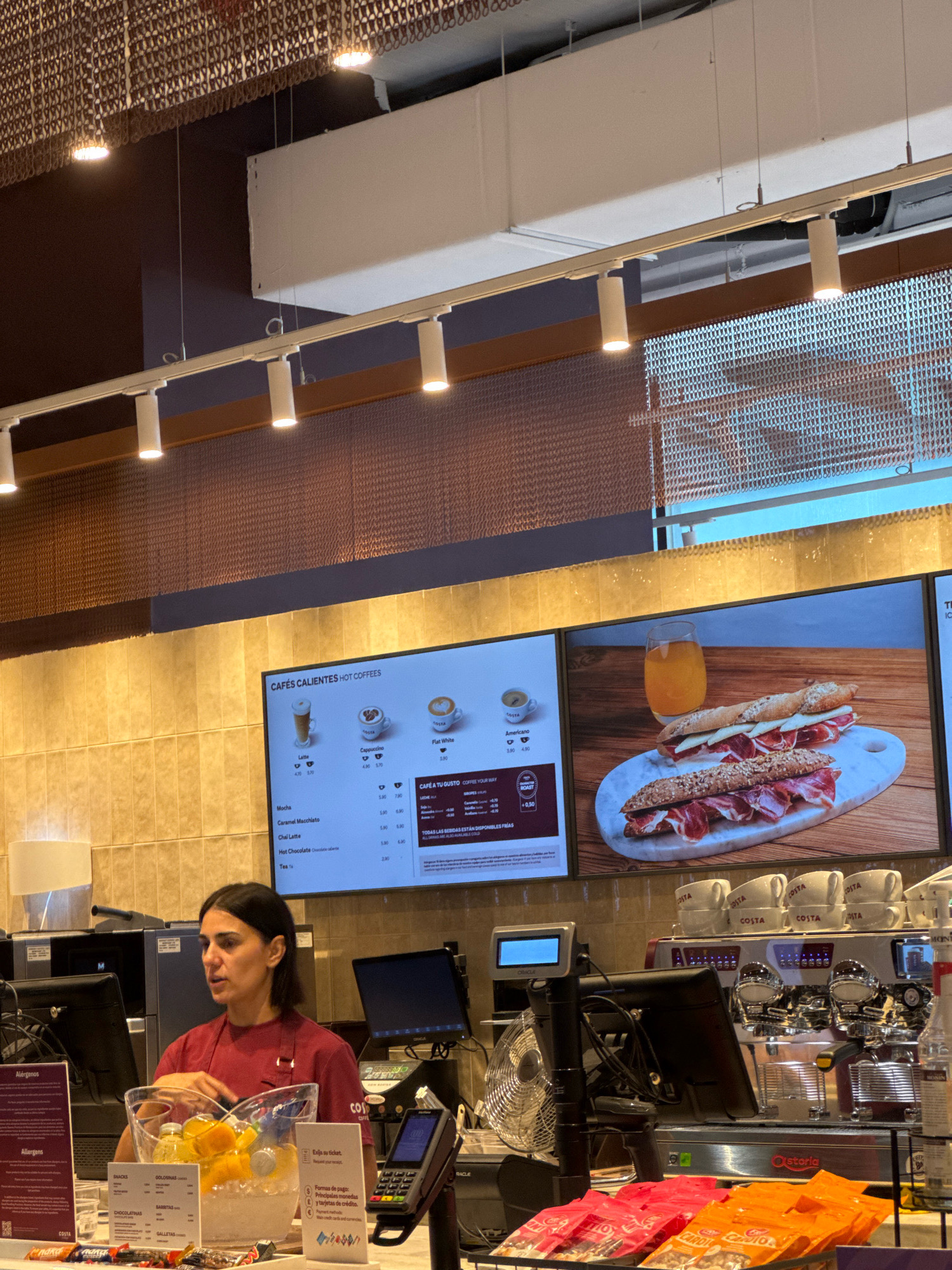 The image depicts the interior of a Costa Coffee shop in Palma de Mallorca, Spain. A female employee, wearing a red t-shirt and apron, is standing behind a counter, interacting with customers. She is positioned to the left of the frame, gesturing with her right hand towards a clear plastic container filled with ice and sliced oranges, suggesting she is preparing or serving a beverage. In front of her are various food items, including packaged snacks and drinks, and a glass bowl with more orange slices.
Behind the employee, a digital menu board displays hot coffee options with prices and images. To the right of the menu board, a large screen shows a close-up image of two sandwiches on a marble board, accompanied by a glass of orange juice. Further to the right, a commercial espresso machine is visible, with a stack of Costa Coffee branded cups on top. A point-of-sale system, including a credit card reader and a small display screen, is also present on the counter.
The environment is well-lit, with artificial lights illuminating the counter area and the digital displays. The background features a tiled wall and a decorative metallic ceiling. The overall scene conveys a typical commercial setting within a coffee shop.