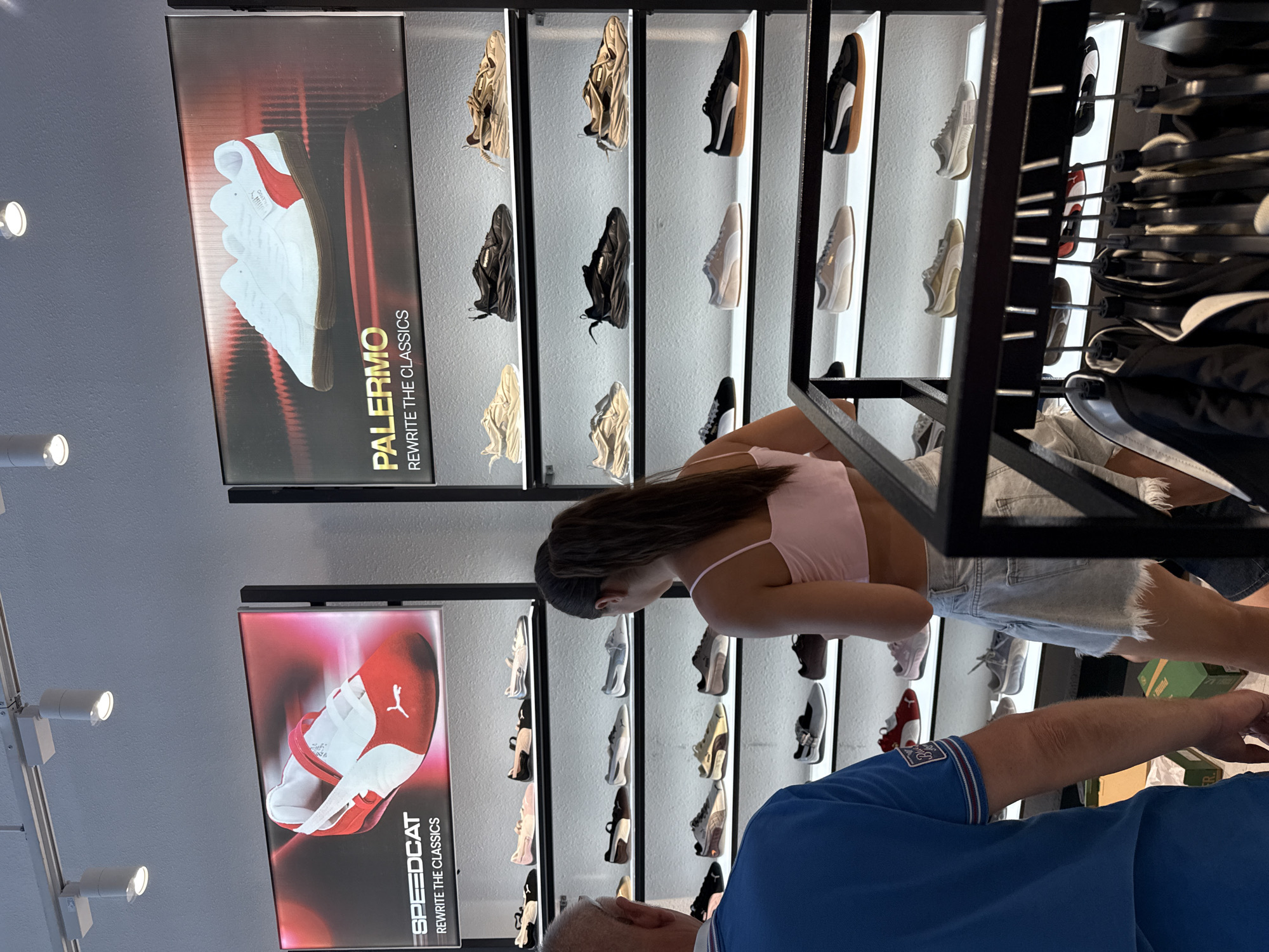 The image depicts the interior of a Puma shoe store in Nha Trang Ward, Vietnam. A man in a blue polo shirt and a woman in a pink crop top and denim skirt are browsing the merchandise. The store features illuminated display boards showcasing Puma "Speedcat" and "Palermo" shoes. Shelves behind the shoppers are stocked with various Puma sneakers. In the foreground, a rack of clothing hangs, suggesting a retail environment. The lighting is bright, typical of a commercial establishment.