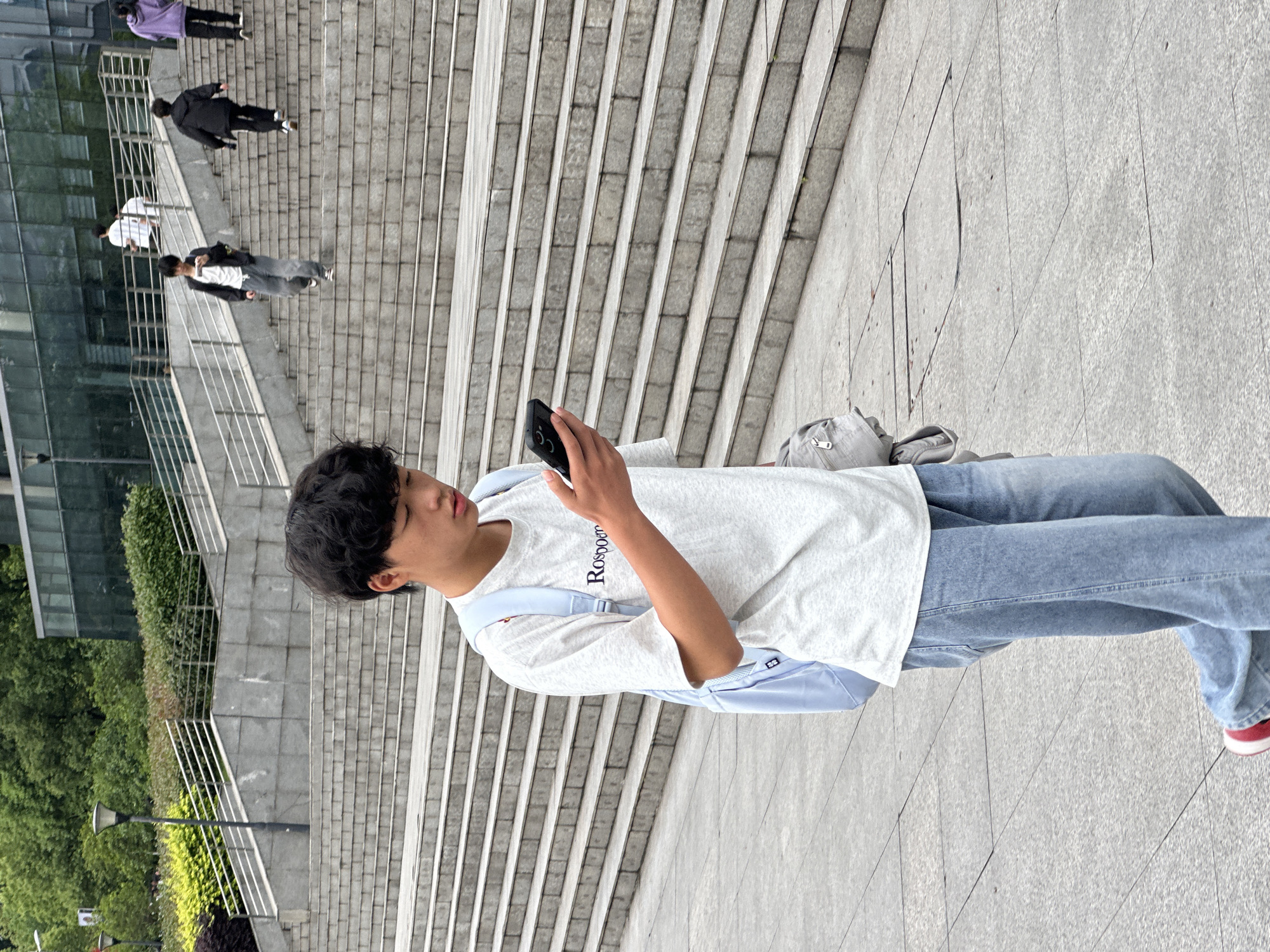 A young man stands on a concrete surface with a light gray color, possibly a sidewalk or walkway. He is holding a smartphone in his right hand and appears to be looking at it. The man is wearing a white t-shirt with the name "Roskoon" on it and light blue jeans, with red and white sneakers. He has short, dark hair. A light blue backpack is slung over his shoulder.
In the background, several people are walking up or down a wide set of concrete stairs. There are metal handrails alongside the stairs. The surrounding environment includes green trees and bushes, as well as a building with glass windows. Based on available information, the location is Jiangjin, China.