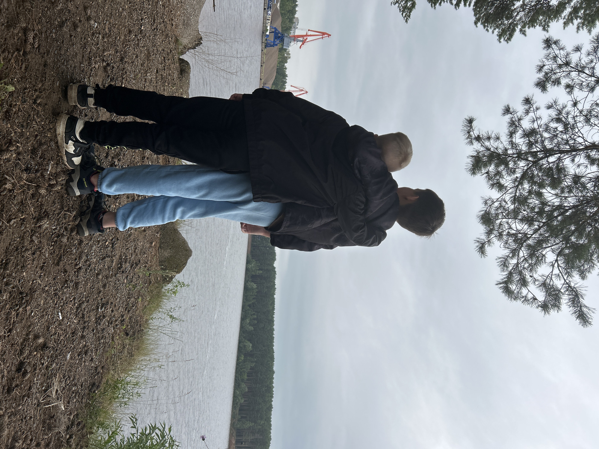 Two people are standing on a gravelly shore overlooking a body of water. The person in front has dark hair and is wearing a black jacket and light blue pants. They are facing away from the camera, looking towards the water. Behind them, another person with lighter hair is embracing them from behind. This person is wearing a black jacket. Both individuals are wearing athletic shoes. The scene appears to be a natural outdoor setting, possibly a lake or river, with a dense forest on the far bank. In the background, industrial cranes and piles of sand or gravel suggest a port or construction area. The sky is overcast, indicating cloudy weather. The time of day is indeterminate due to the cloud cover. The overall mood seems relaxed and peaceful. There is no visible text in the image.