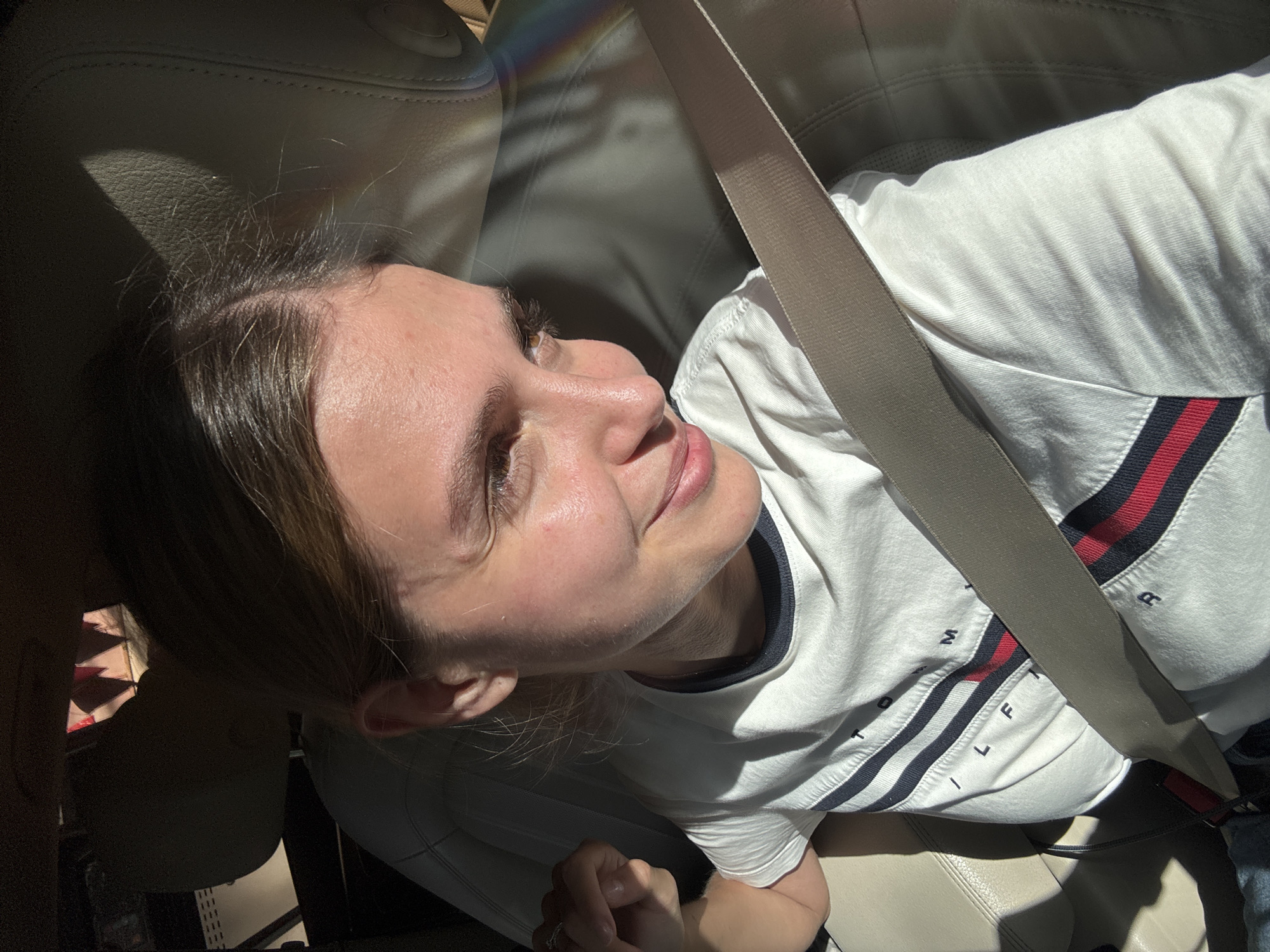A young woman is shown from an angled, overhead perspective, secured in a car seat with a beige seatbelt across her torso. She is wearing a white t-shirt with dark blue and red stripes and what appears to be the word "HILFIGER" printed on it. The interior of the car is visible, with beige leather seats. Sunlight is streaming into the car, creating strong contrasts and a rainbow-like effect on the upper left of the image.