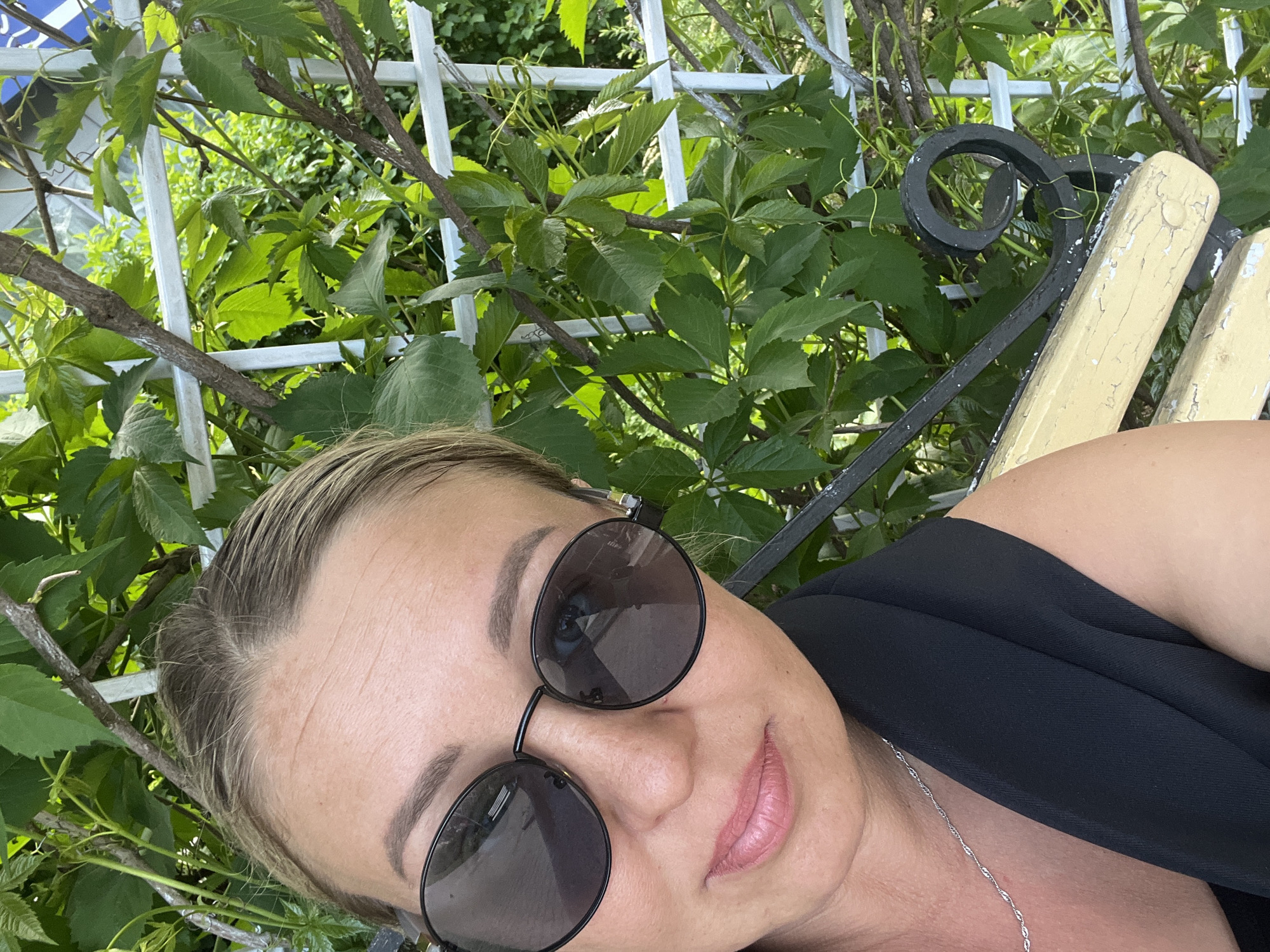 A woman wearing sunglasses and a black top is posing for a selfie. She is sitting on a yellow and black bench. Behind her is a white trellis covered in green vines. The location appears to be an outdoor area, possibly a park or garden. The image does not contain enough information to determine if the location is in the city of Chaikovsky, Russia.
