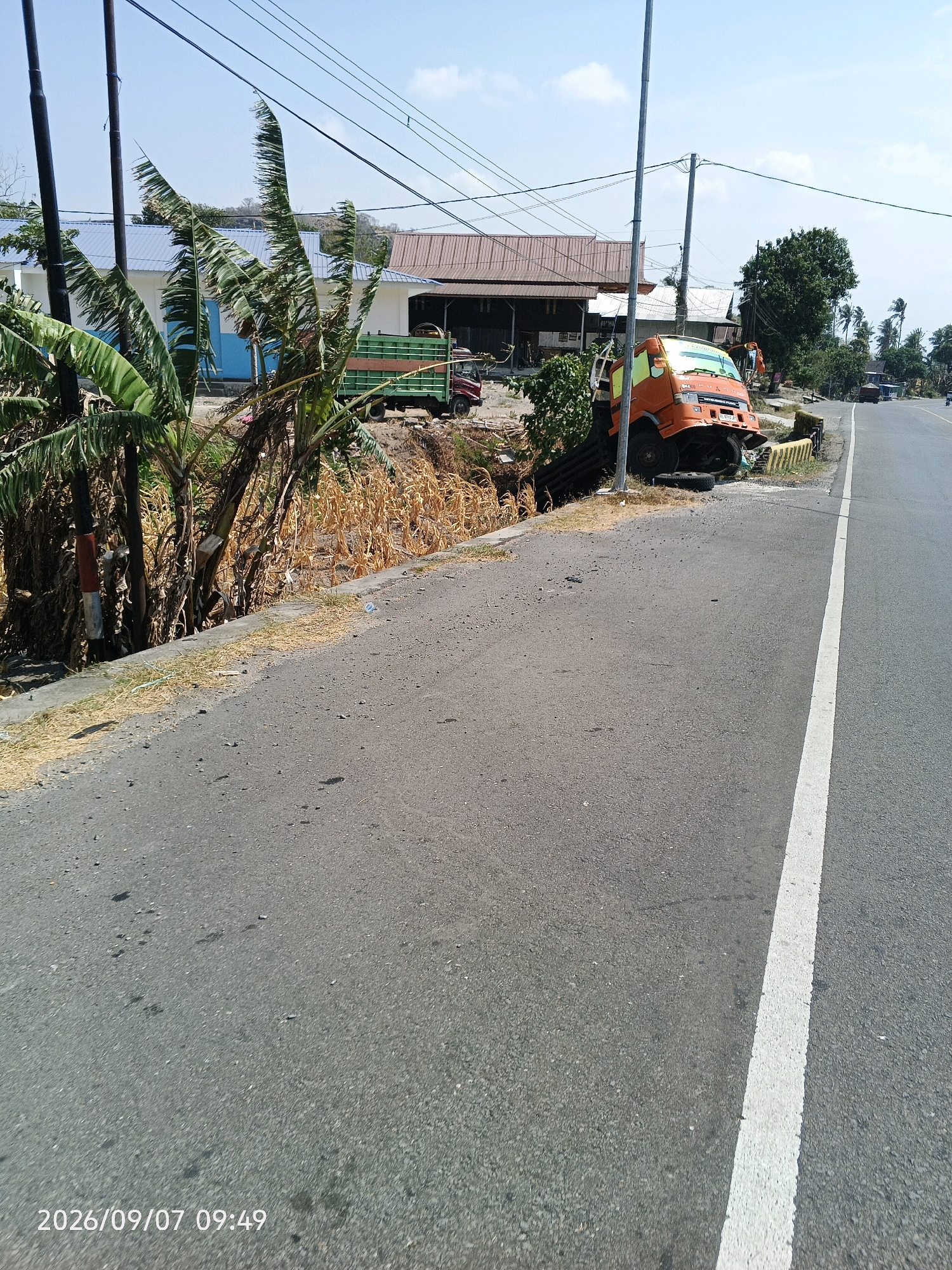 In Lawawoi, Indonesia, an orange and yellow truck has crashed off a road. The truck is severely damaged, with its front end crumpled and its wheels off the ground. It has fallen into a ditch next to the road, which is lined with yellow and white barriers. The surrounding area is overgrown with vegetation, including banana trees and dry, brown grass. In the background, there are several buildings with corrugated roofs, as well as power lines and utility poles. The sky is clear and blue, suggesting it is a sunny day. The date stamp in the lower left corner reads "2026/09/07 09:49".