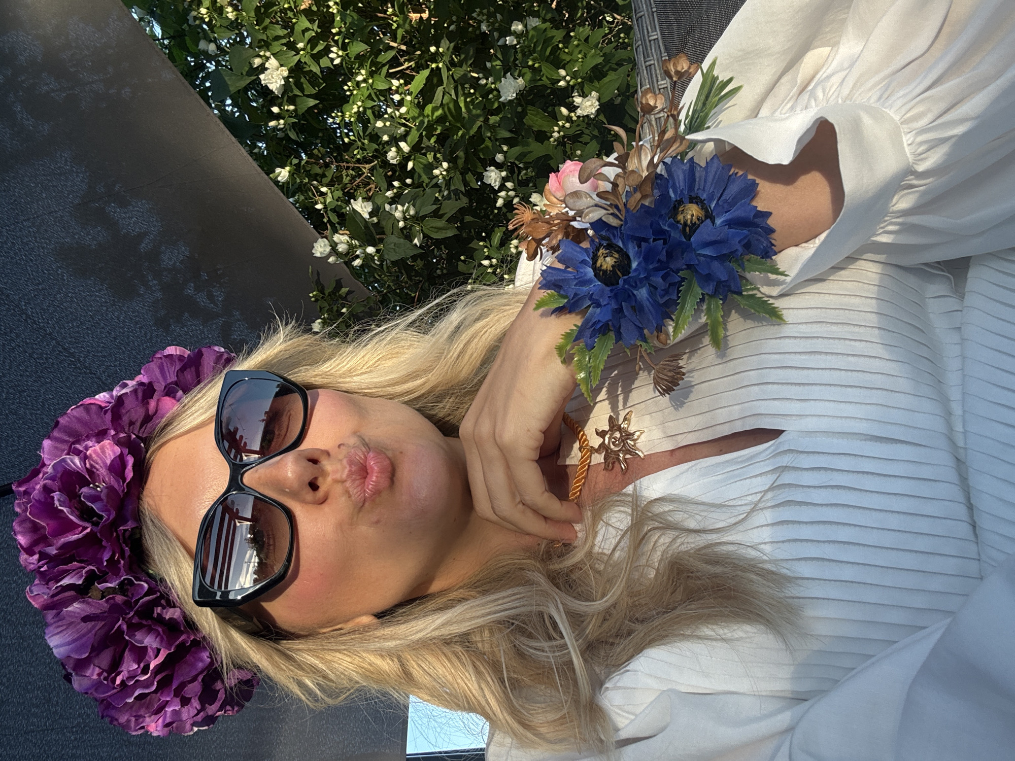 This media file is a selfie taken outdoors, likely during the day, as indicated by the bright lighting and shadows. The primary subject is a woman with blonde hair, wearing large black sunglasses and a vibrant purple flower crown. She is pouting with her lips pursed. On her left side, she is adorned with a small bouquet of blue and pink artificial flowers, possibly a boutonniere or part of her attire. She is wearing a white, loosely-fitting top with puffy sleeves and a pleated texture on the bodice. Her hand is raised to her chest, touching the flower bouquet.
Behind her, lush green foliage with small white flowers is visible, suggesting a garden or natural setting. A dark, textured surface, possibly a piece of furniture or a wall, occupies the upper left portion of the frame. The overall scene evokes a feeling of celebration or a whimsical outdoor event, possibly in a warm climate given the abundant greenery and bright light. The woman's expression and attire suggest a lighthearted and joyful mood. No visible text is present in the image. The location context provided is Carnikava, Latvia, which aligns with a natural, outdoor setting with blooming flora.
