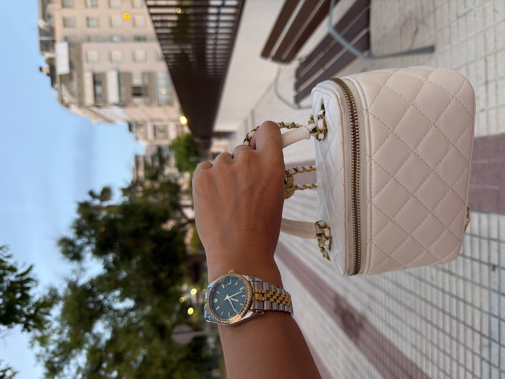 An outdoor medium shot shows a person's right arm and wrist, wearing a two-tone gold and silver watch with a green face, holding a small white quilted handbag with a gold chain strap and zipper. The background shows a paved walkway with a reddish stripe, trees, a metal bench, and buildings. The sky is a pale blue.