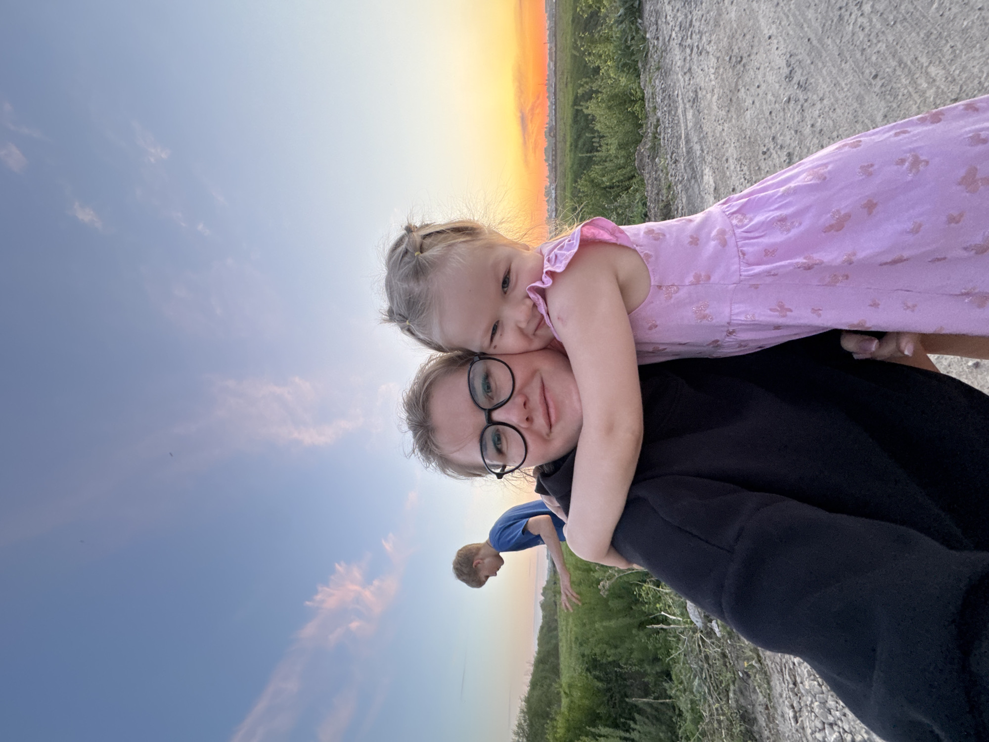 A woman wearing glasses and a black jacket is holding a young girl in a pink dress. The girl is hugging the woman. A young boy in a blue shirt stands to the left, near the horizon. The location is Горный, Russia, at sunset. A gravel road and green vegetation are visible. The sky is transitioning from blue to orange.