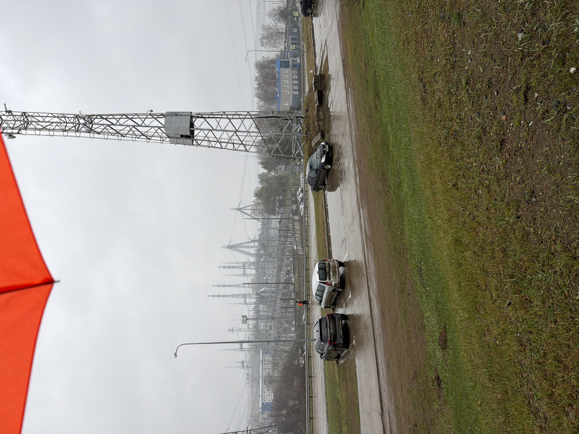 The image depicts an overcast and wet day in Chaikovsky, Russia, with a slightly tilted perspective. In the immediate foreground on the left, a vibrant orange umbrella or canopy is partially visible. The right side of the foreground features a grassy verge with patches of bare earth.
The midground is dominated by a dark, wet asphalt road with visible puddles reflecting the gloomy sky. Several vehicles are parked along the roadside: a black sedan with license plate R366TA 199, a white utility vehicle or SUV displaying a "Coca-Cola" logo on its rear door, and a dark SUV with license plate X713BP 199. A person wearing a high-visibility orange vest stands near the rear of the white vehicle, appearing to be working or inspecting something. A large, intricate metal lattice structure, resembling an electricity pylon, stands prominently on the left side of the midground.
In the background, a vast electrical substation unfolds, characterized by numerous tall pylons, transformers, and a complex network of power lines, all silhouetted against the uniformly grey sky. Further in the distance, buildings and leafless trees are faintly visible through a general atmospheric haze, suggesting an industrial or peripheral urban setting. The wet road surfaces and heavy cloud cover indicate recent rainfall or ongoing damp conditions, creating a somber and utilitarian atmosphere.