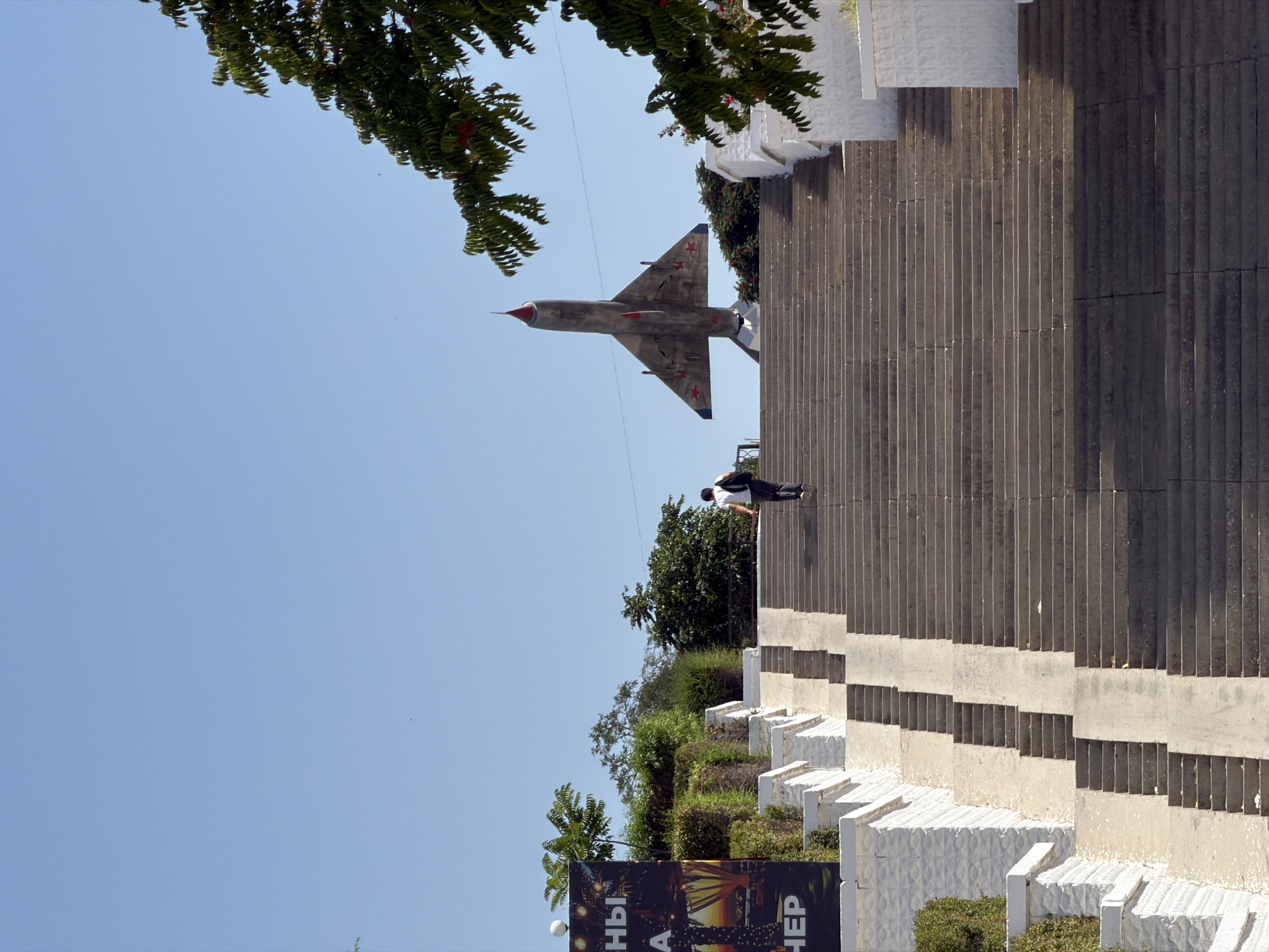 A lone individual, seen from behind, ascends a wide, sun-drenched stone staircase. The person, wearing a dark backpack, is positioned in the upper right quadrant of the frame. At the top of the staircase, a weathered, grey jet fighter monument stands proudly against a clear blue sky. The monument is adorned with red stars on its wings and fuselage, and its nose cone is painted red. The staircase is flanked by low, white-painted walls and lush green foliage. To the left of the staircase, a large poster with Cyrillic lettering is partially visible, featuring colorful graphics and the word "НЕР". The image appears to be taken in the city of Aktau, Kazakhstan, likely at a memorial site dedicated to aviation. The bright sunlight casts distinct shadows on the steps, indicating it is daytime.