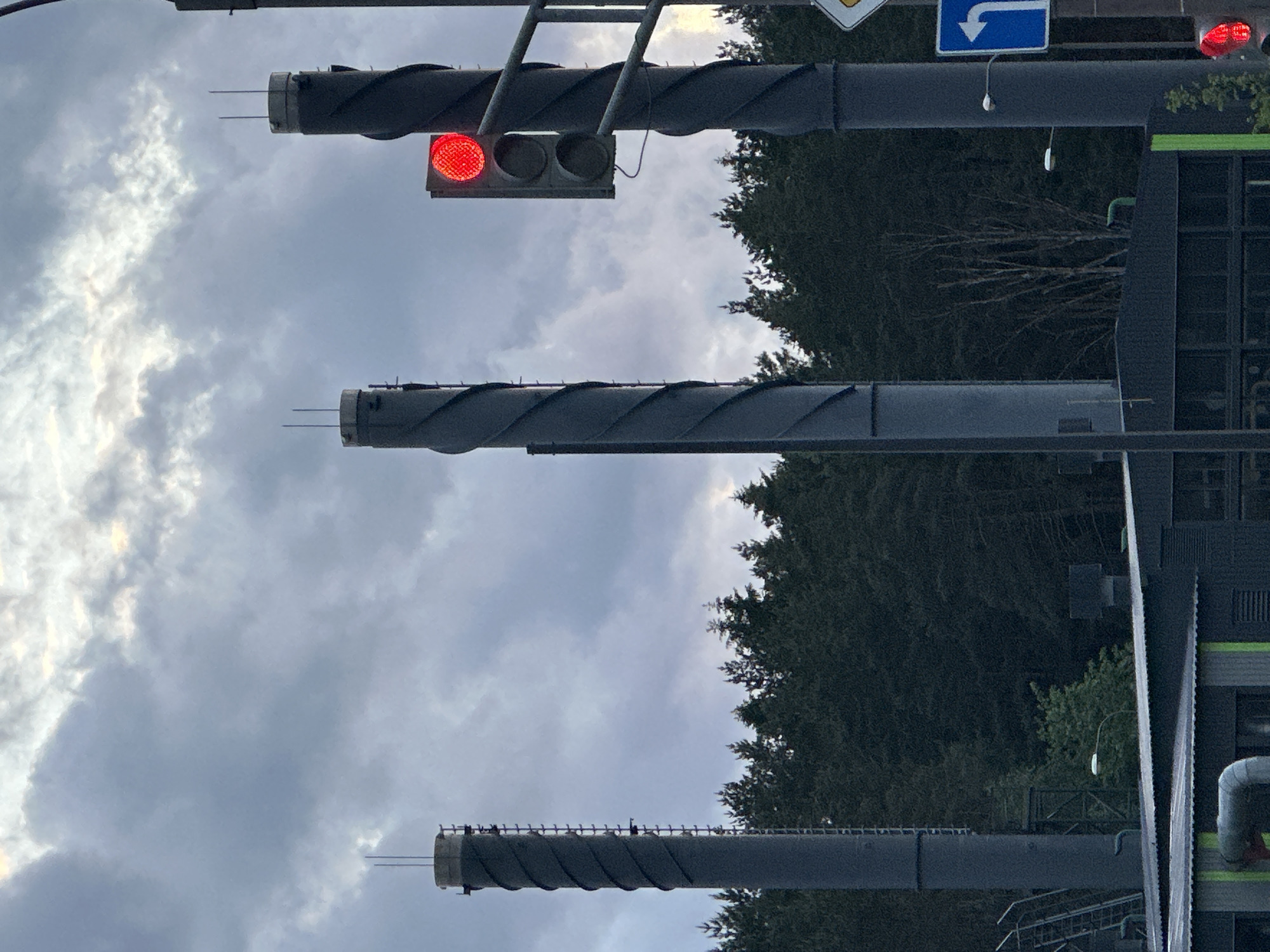 The image shows three tall, gray industrial smokestacks against a cloudy sky and some trees. The smokestacks have a unique, twisted exterior design. The top smokestack has a traffic light with a lit red light. A blue right turn sign is also visible. Industrial buildings and infrastructure elements are visible to the right of the image. The location is likely in or near Бараўлянскі сельскі Савет, Belarus, considering the industrial setting and the smokestacks. The scene appears still and empty of people.
