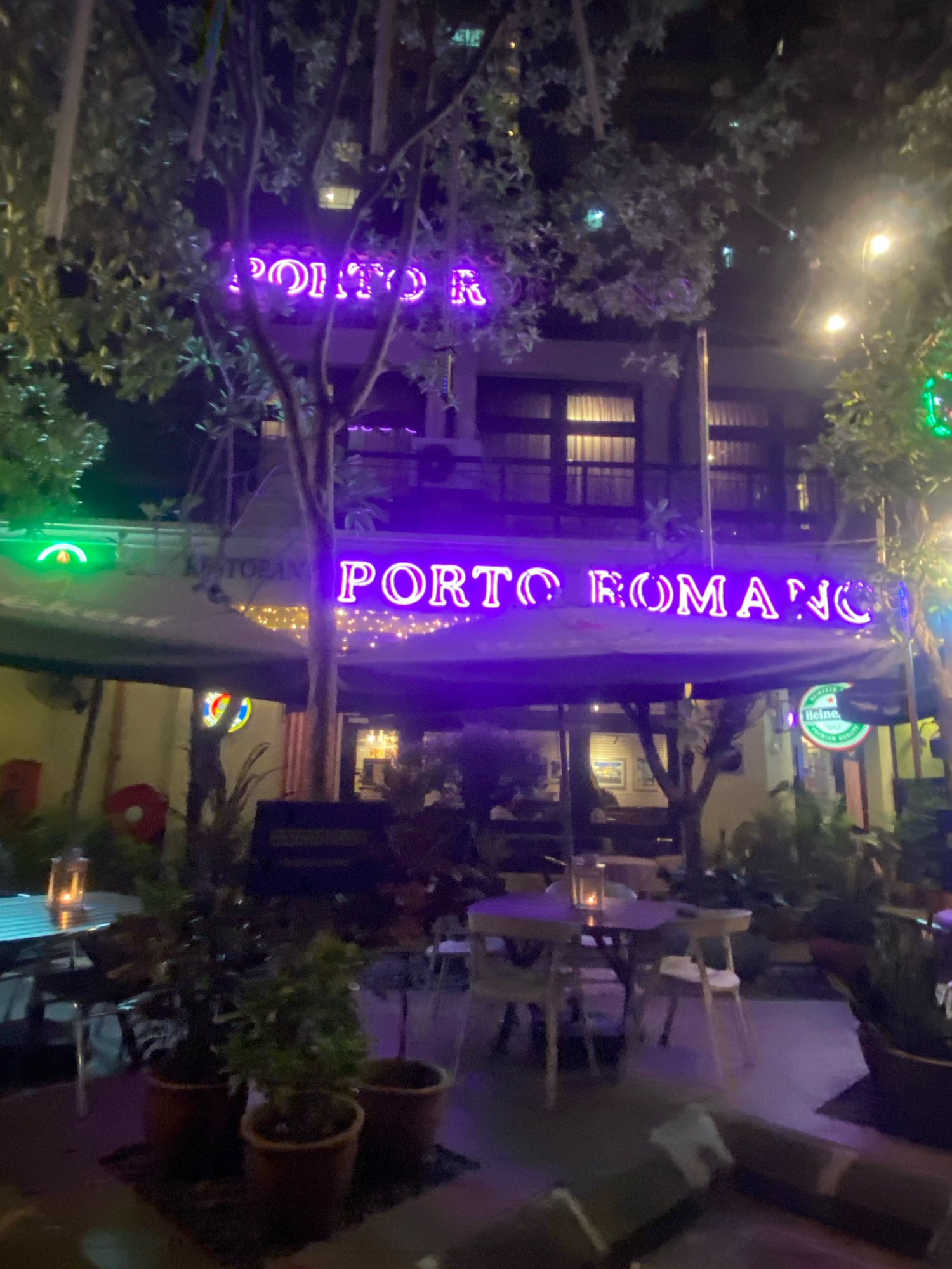 The image depicts the exterior of "Restoran Porto Romano" at night in Kuala Lumpur, Malaysia. A prominent neon sign in bright purple spells out "PORTO ROMANO" across the front of the establishment. The restaurant appears to have an outdoor seating area with tables and chairs, some of which are visible in the foreground and midground. Trees with lush foliage are interspersed with the seating, and some are illuminated by string lights. The overall ambiance is that of a lively, illuminated dining spot at night, with a focus on the glowing neon signage.